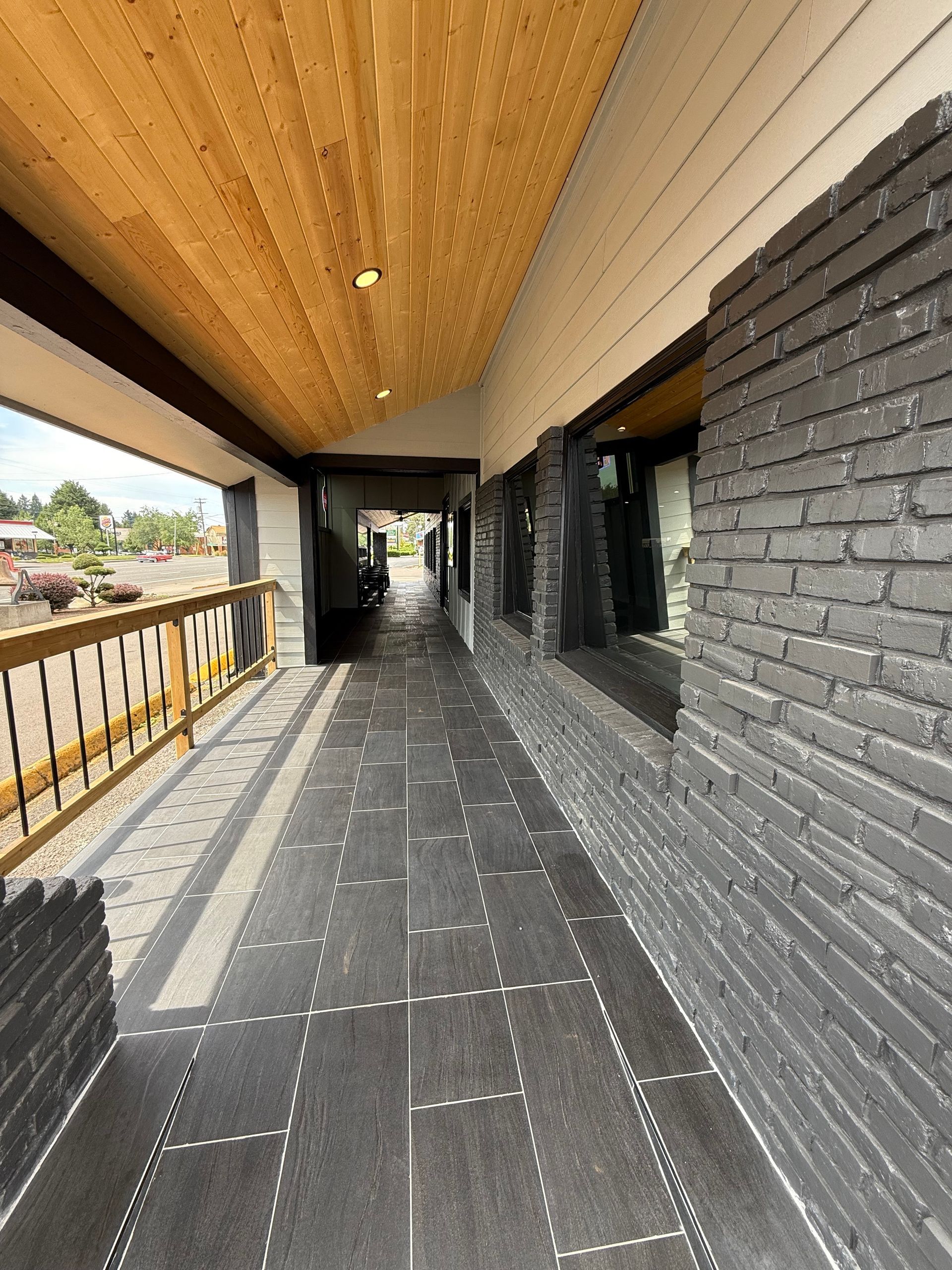 Covered walkway with black brick wall, wood ceiling, and metal railing overlooking street.