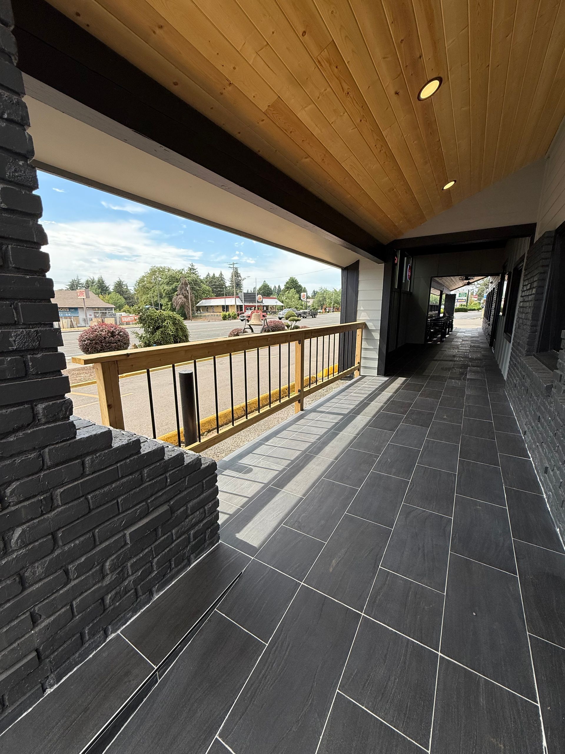 A covered outdoor walkway with black tiled floor, brick wall, and a view of a sunny landscape.