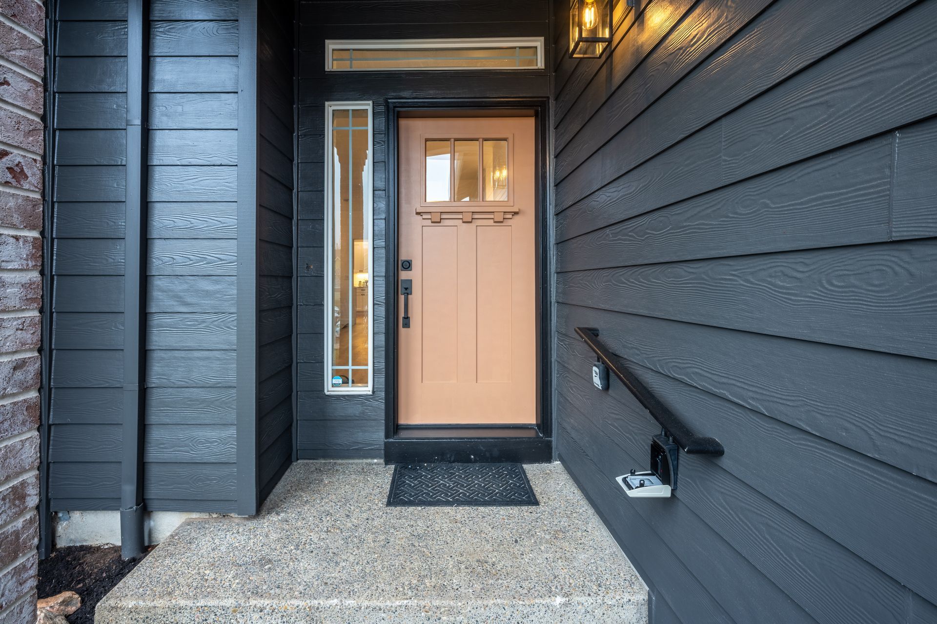 Tan front door with sidelight and black siding on a house.
