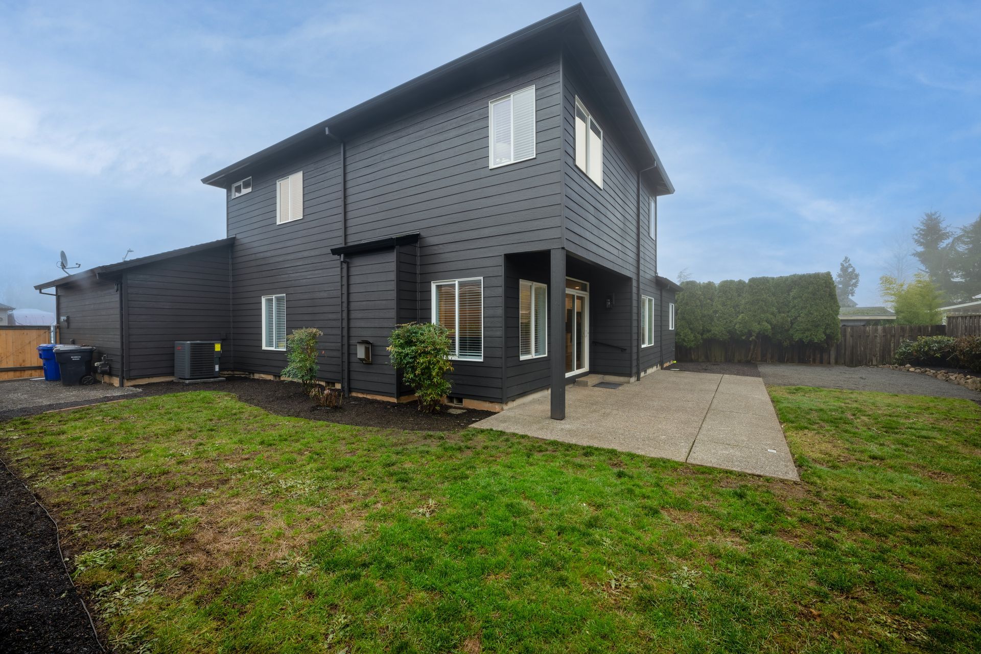 Two-story black house with a concrete patio and grassy backyard under a cloudy sky.