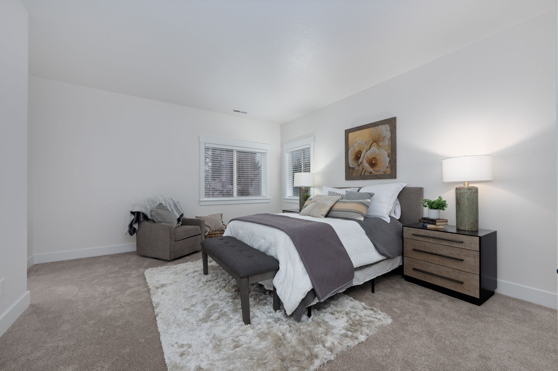Bedroom with neutral tones: bed, nightstand, chair, and rug. Square floral art hangs above bed.