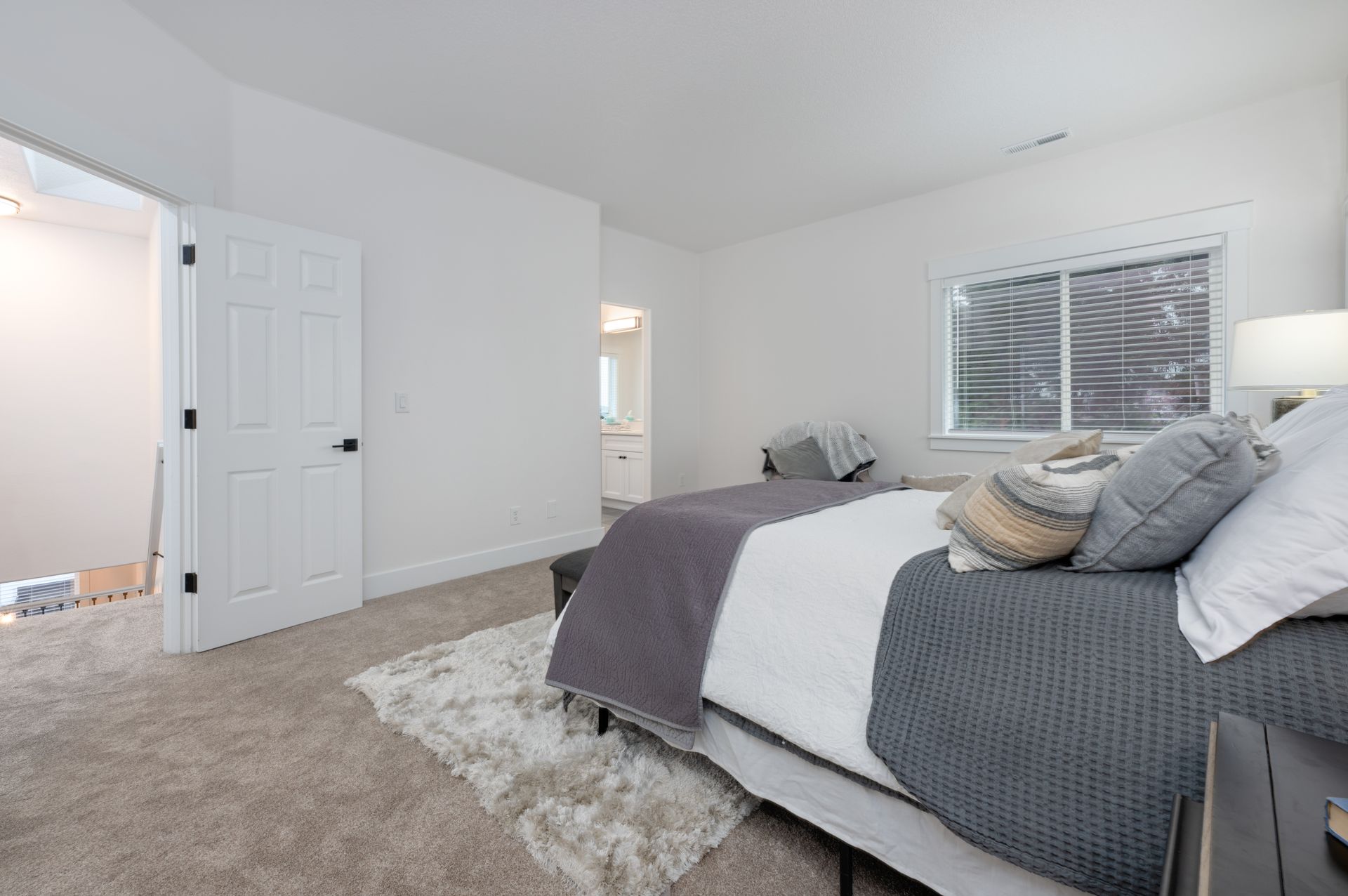 Bedroom with bed, door to bathroom, window, gray and white bedding, neutral carpet.