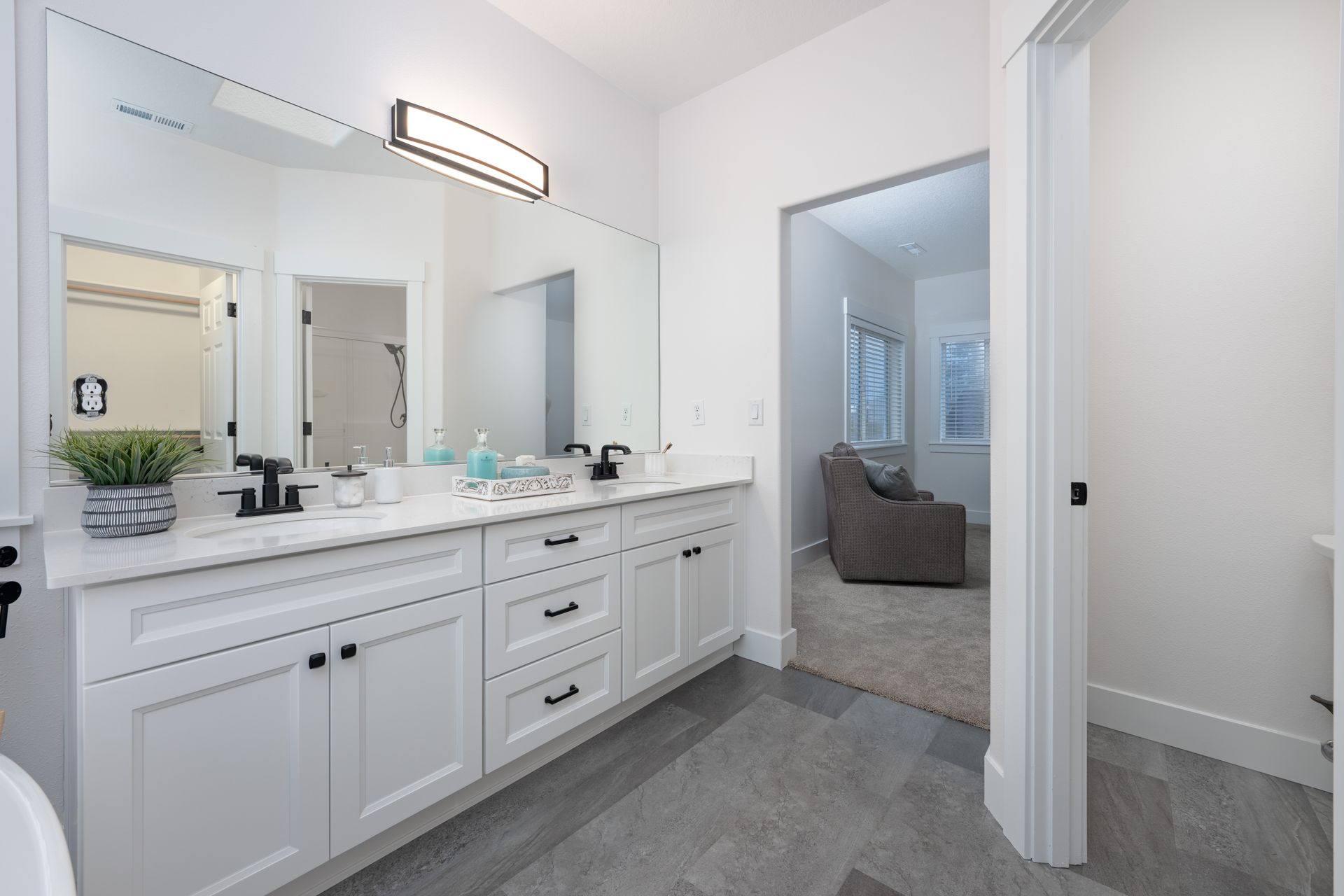 White bathroom with double vanity, large mirror, and doorway to a sitting area with an armchair.