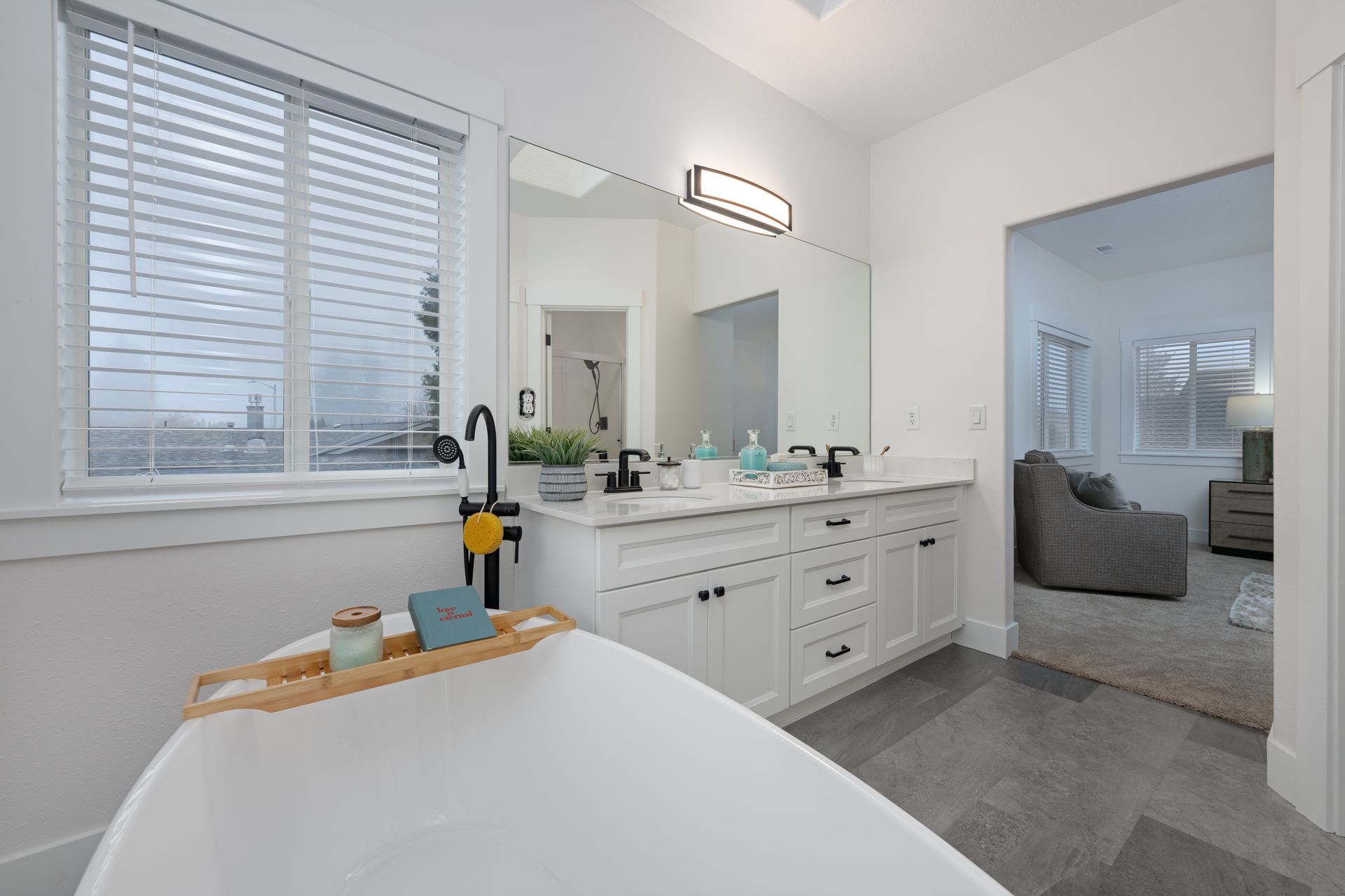 A modern bathroom with a white tub, vanity, and window blinds; a doorway leads to a sitting area.