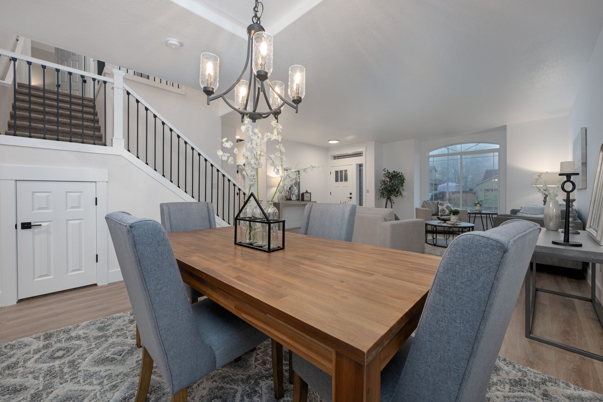 Dining room with wooden table, blue chairs, and chandelier. Staircase and living room in the background.