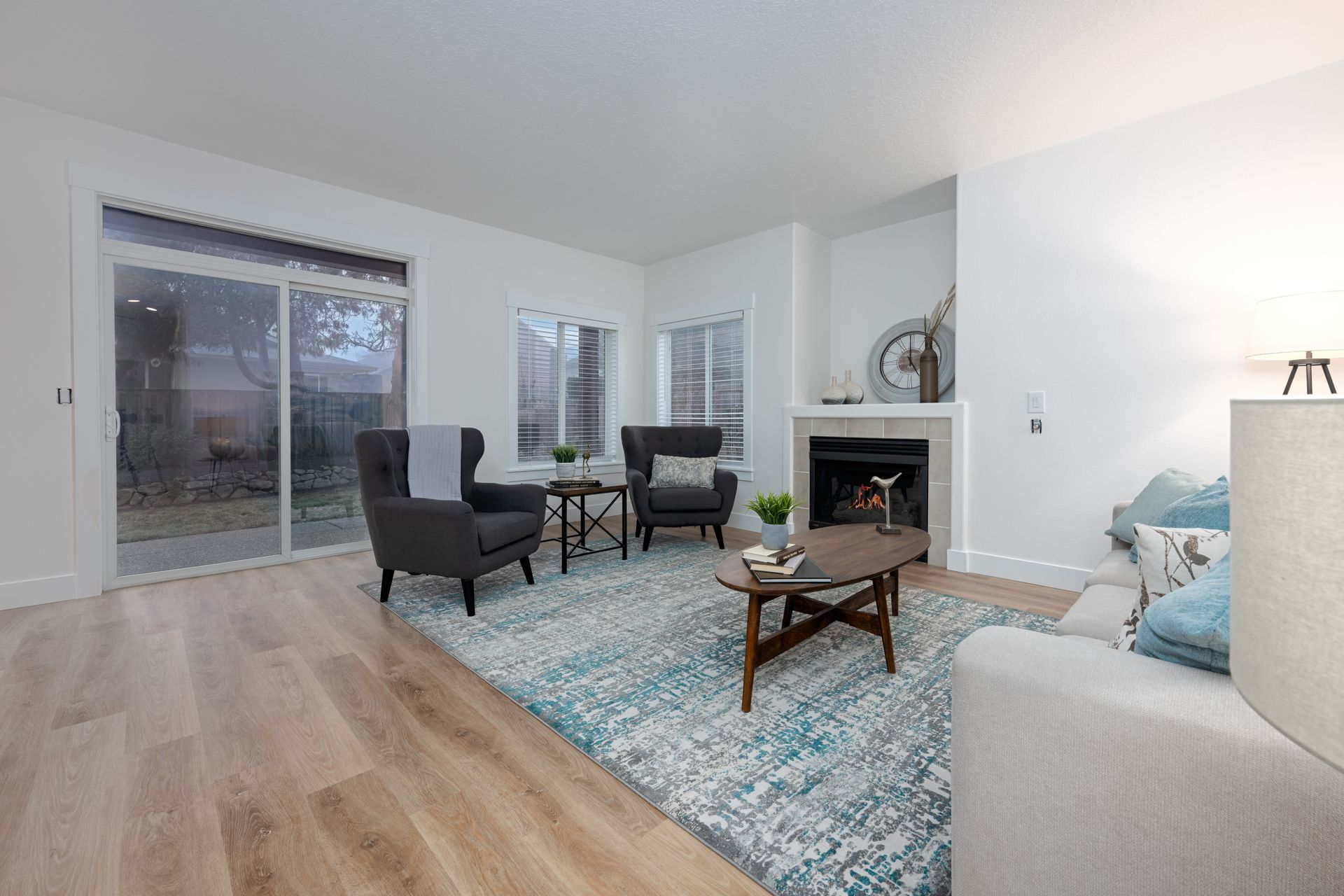 Living room with gray armchairs, fireplace, and blue patterned rug; wooden floor, large window.