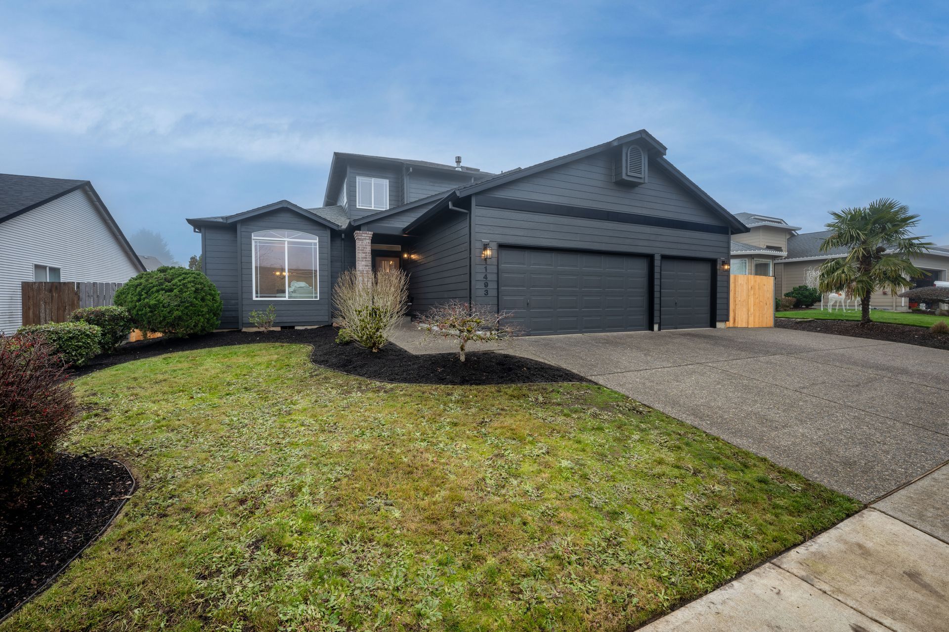 Dark gray house with a two-car garage, green lawn, and a cloudy sky.