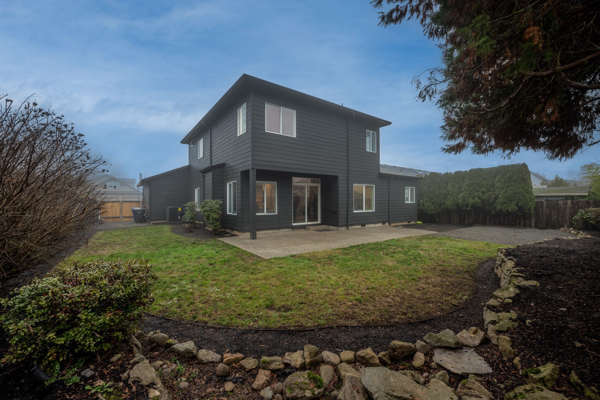 Black two-story house with a backyard patio and green lawn, set under a cloudy sky.