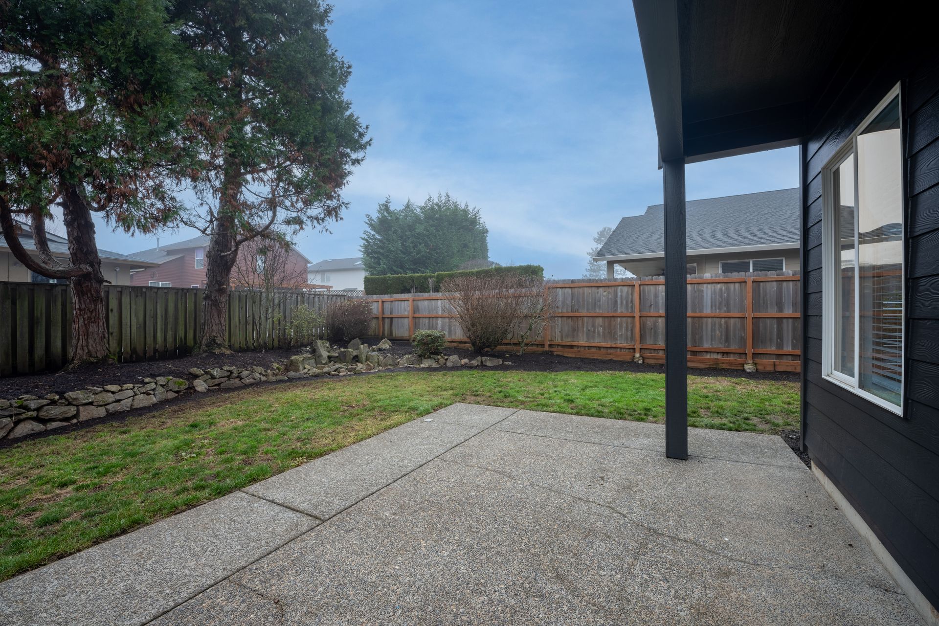 Backyard with concrete patio, grass, wooden fence, and dark exterior siding. Overcast sky.