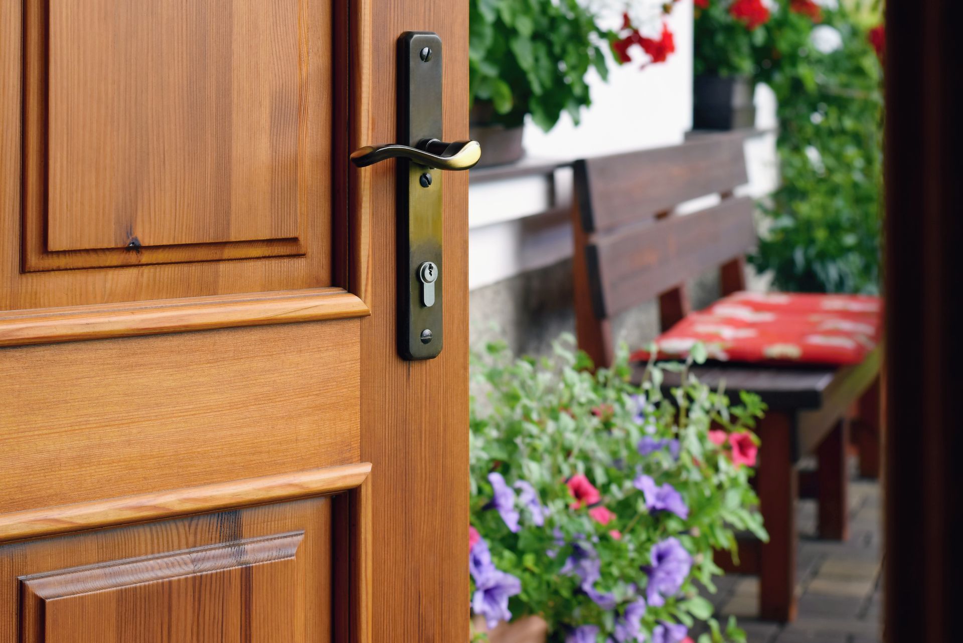 Open wooden front door with black handle beside a flower-filled porch and patio chair
