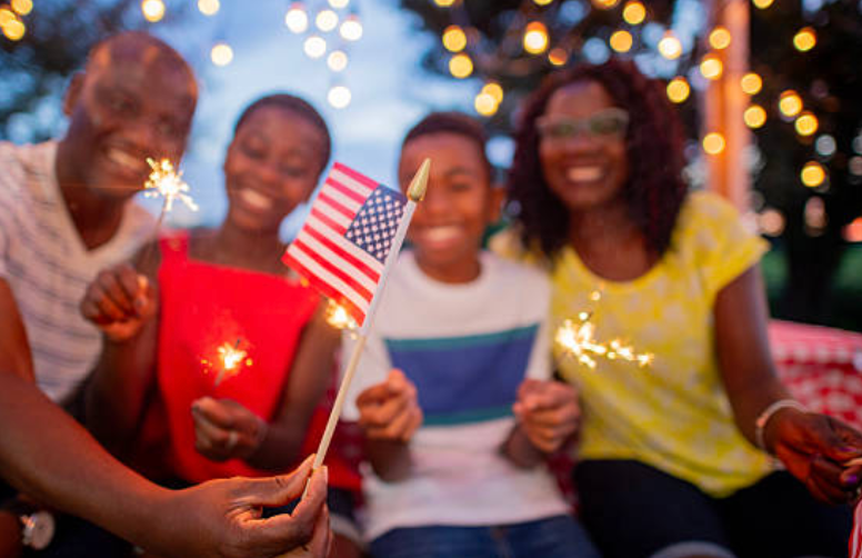 Friends holding candles and a small U.S. flag at a festive evening gathering with colorful lights