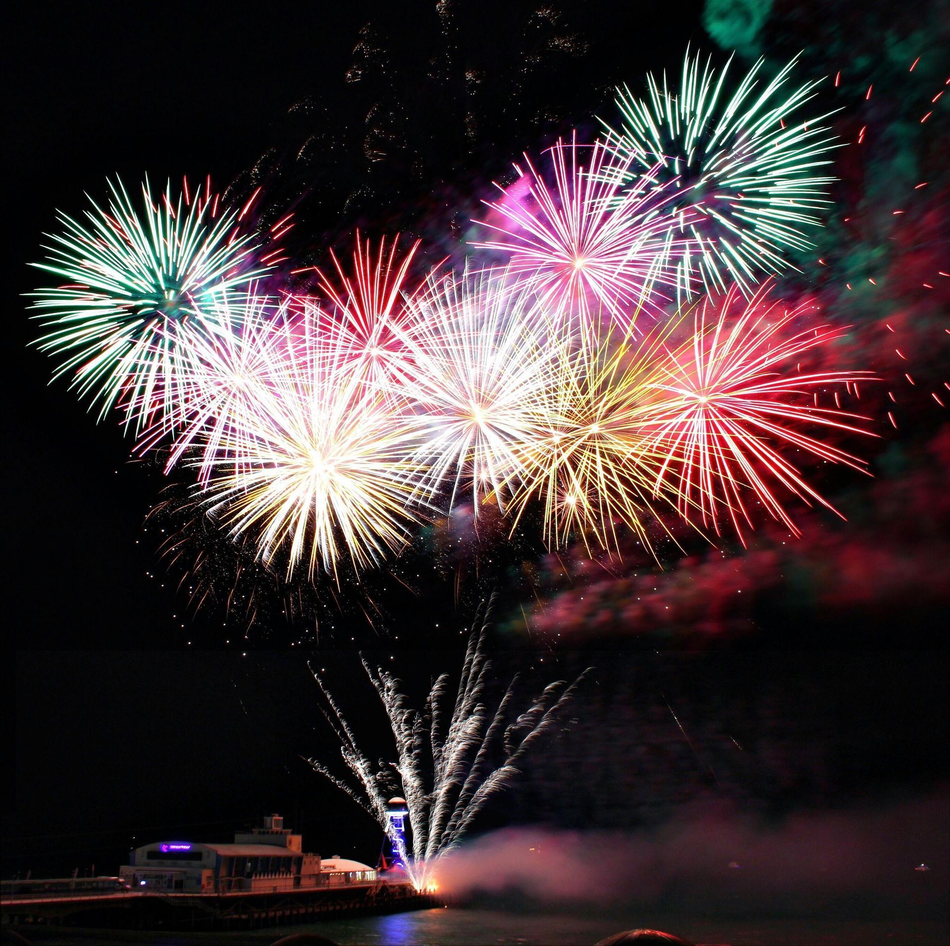 Colorful fireworks bursting over a waterfront at night, with bright reflections near the shore