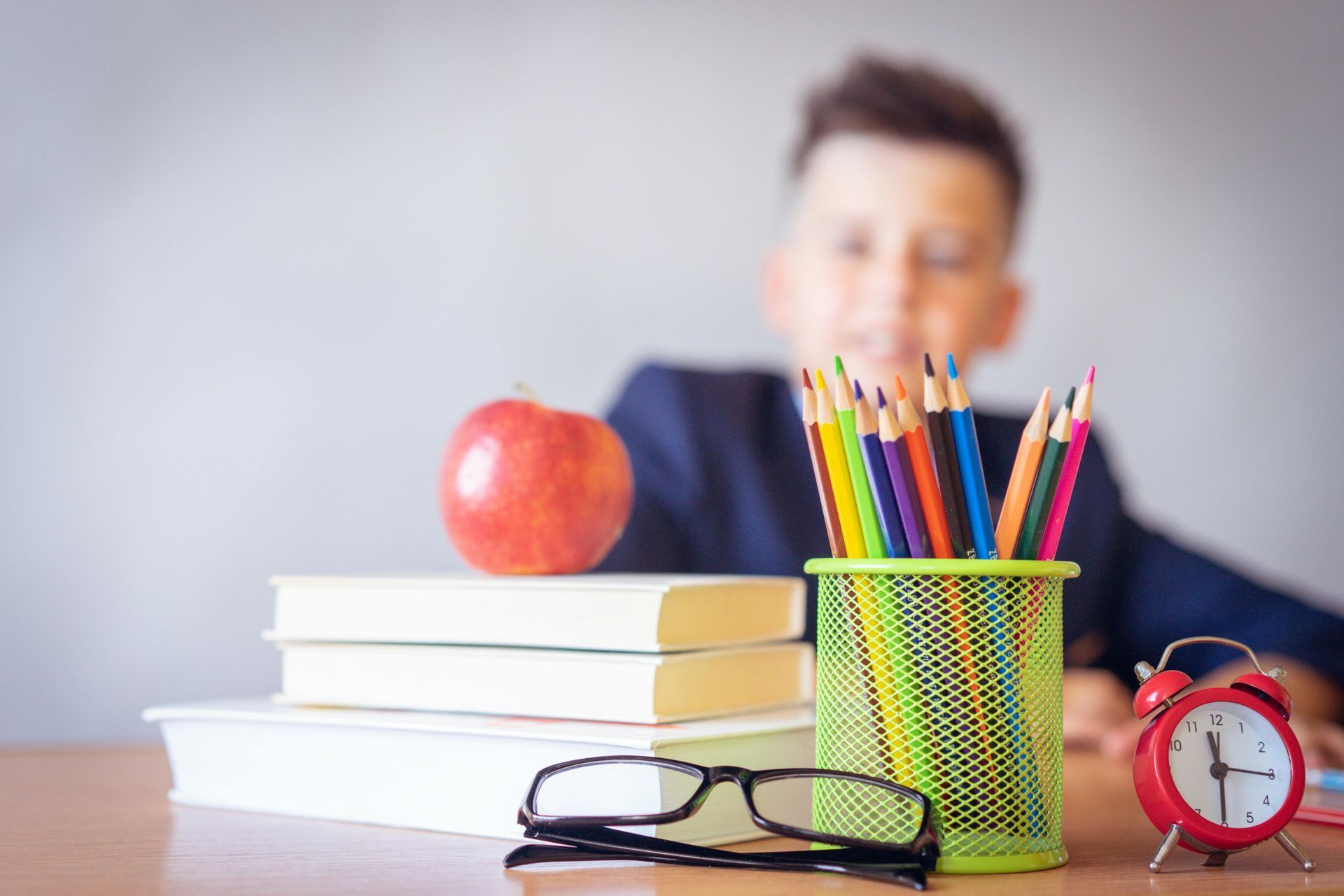 School supplies on a desk with books, pencils, glasses, a red apple, and an alarm clock; blurred person in background