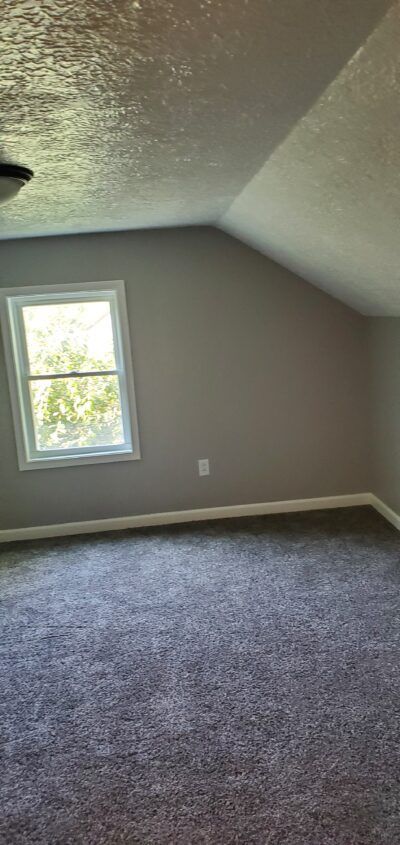 A mostly empty room with gray walls, angled ceiling, window, and gray carpet.