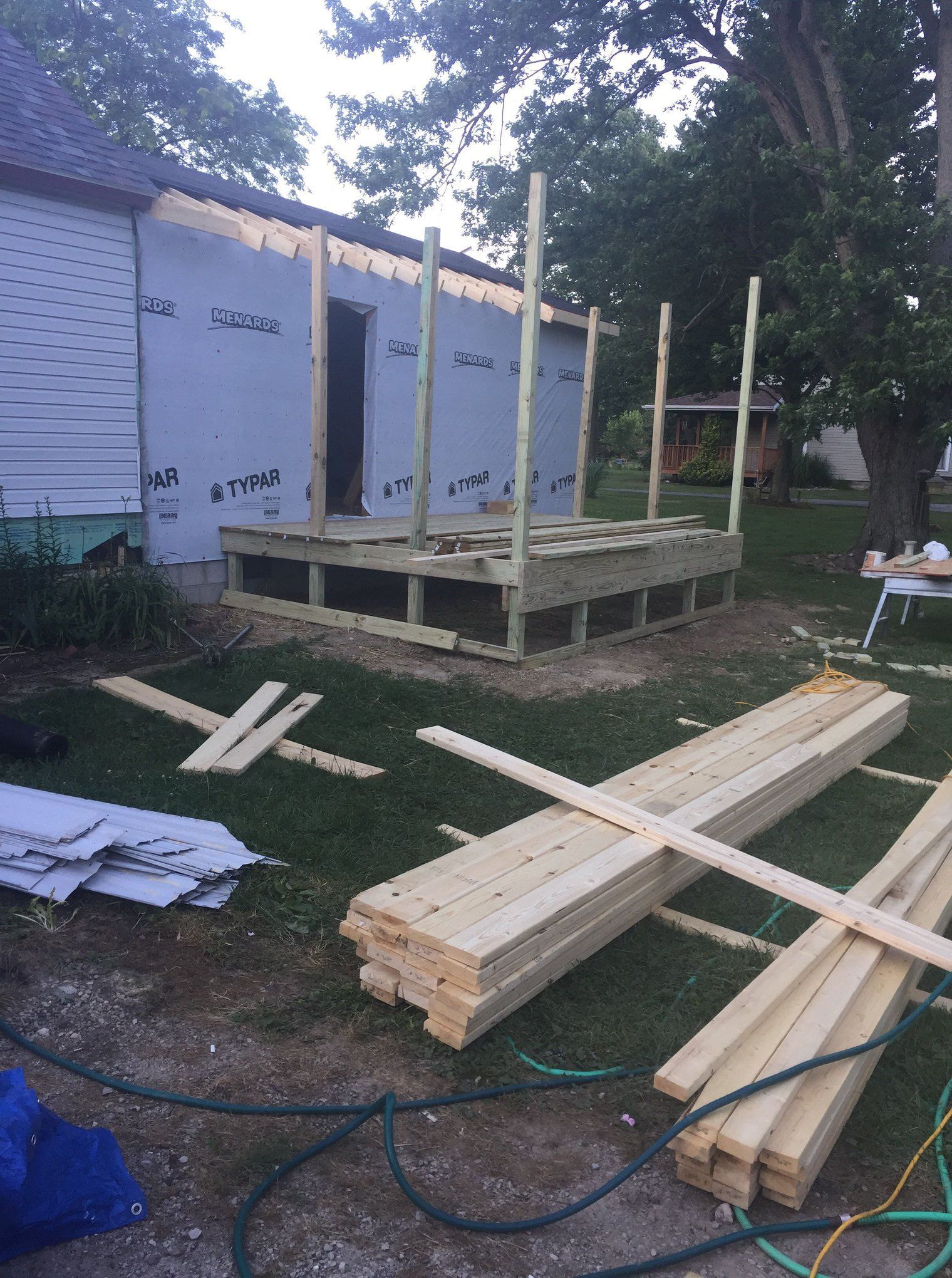 Construction of a wooden deck with upright supports, next to a building with Tyvek wrap. Lumber and tools are scattered on the ground.