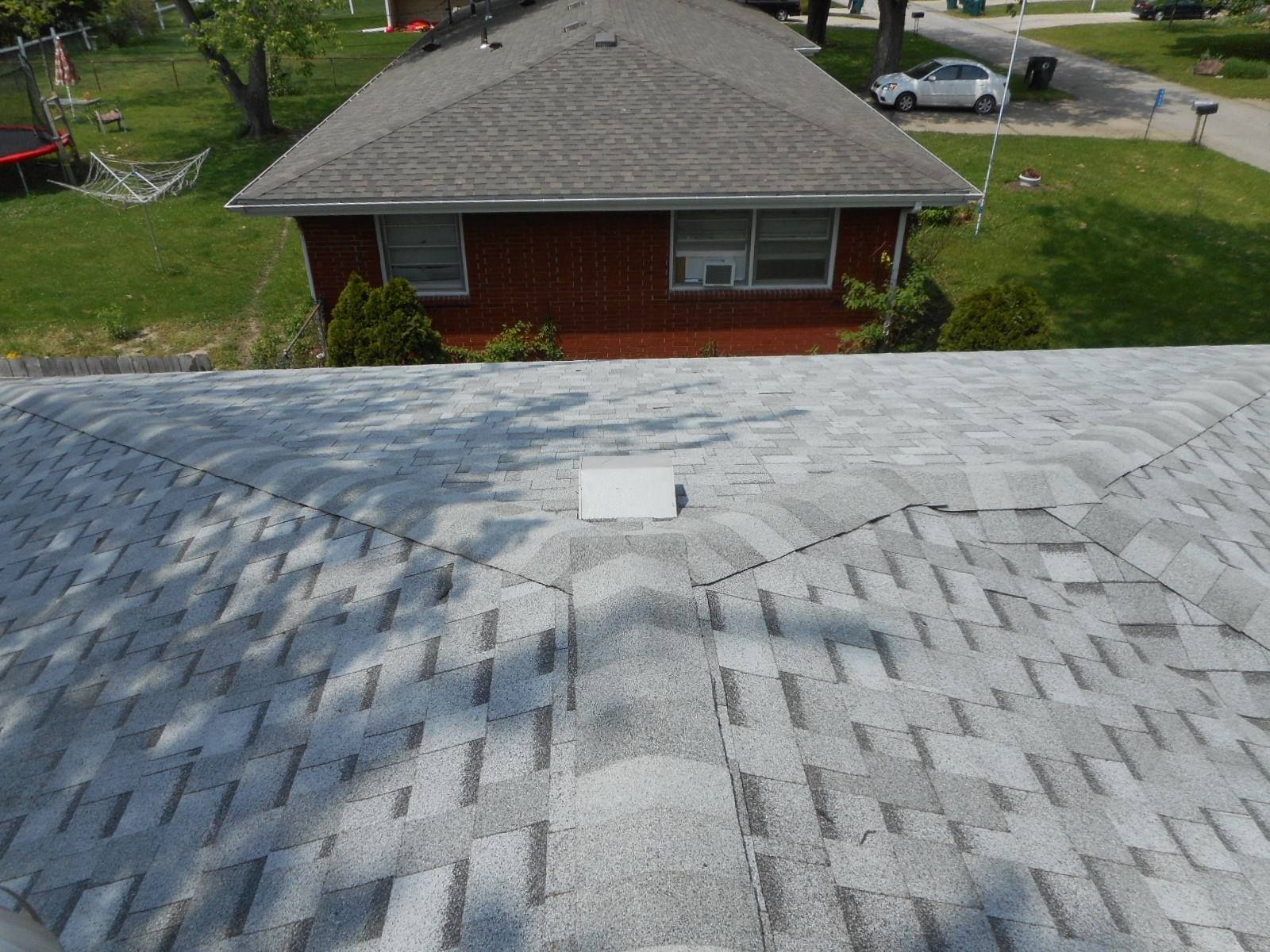 View of a gray shingled roof with a white vent, looking toward a brick house with a gray roof and green yard.