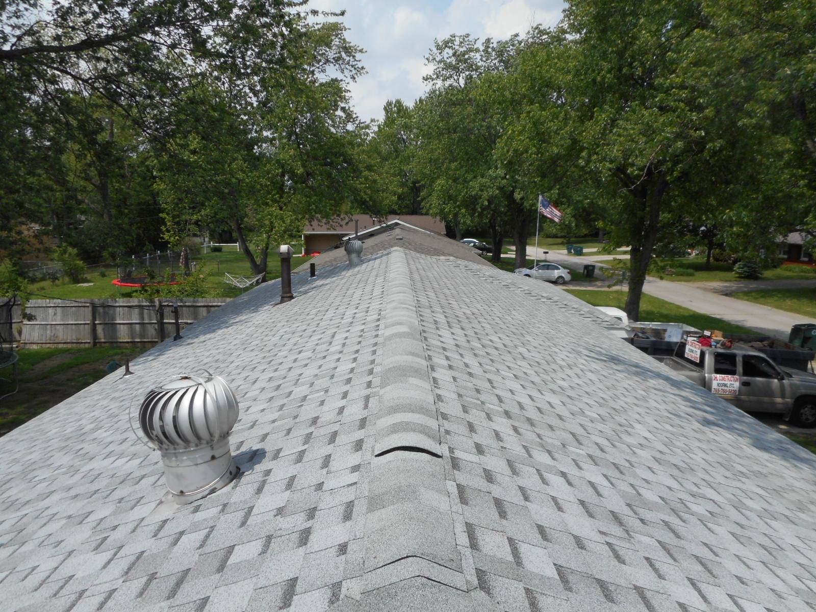 Gray asphalt shingle roof with a ridge vent and a spinning ventilation cap, trees in background.