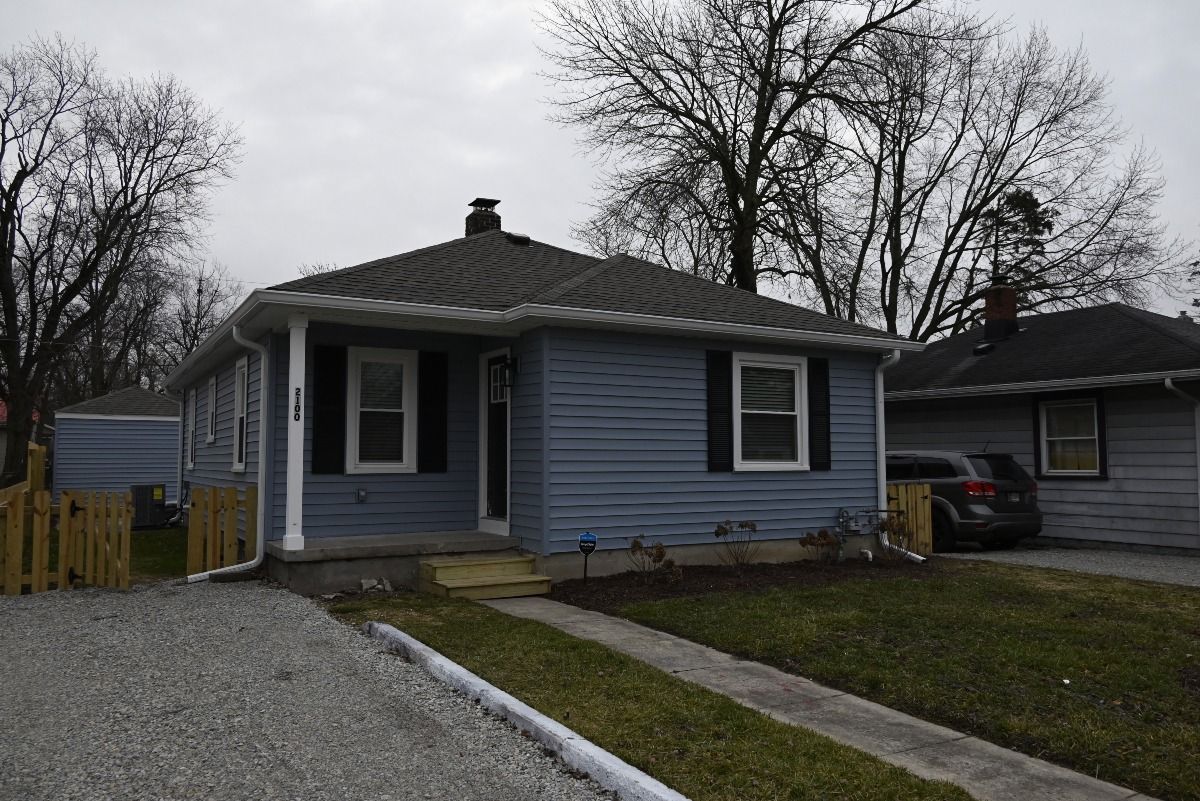 Blue house with black shutters, gray roof, and a gravel driveway.