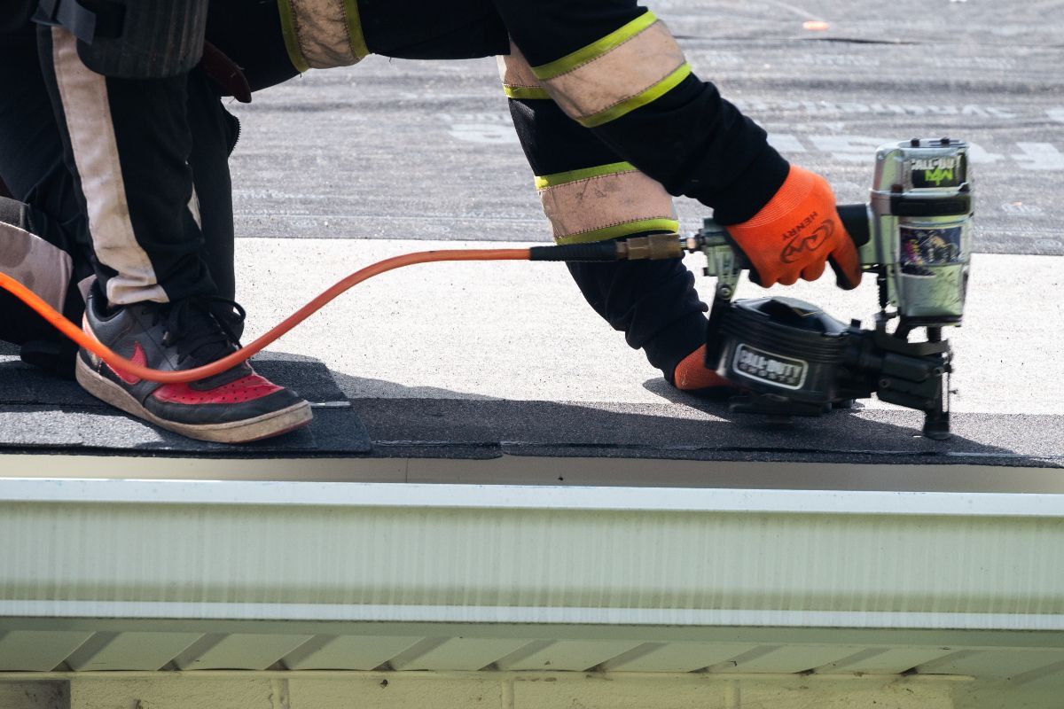 Roofer using a nail gun on a shingle roof, wearing orange gloves and safety gear.