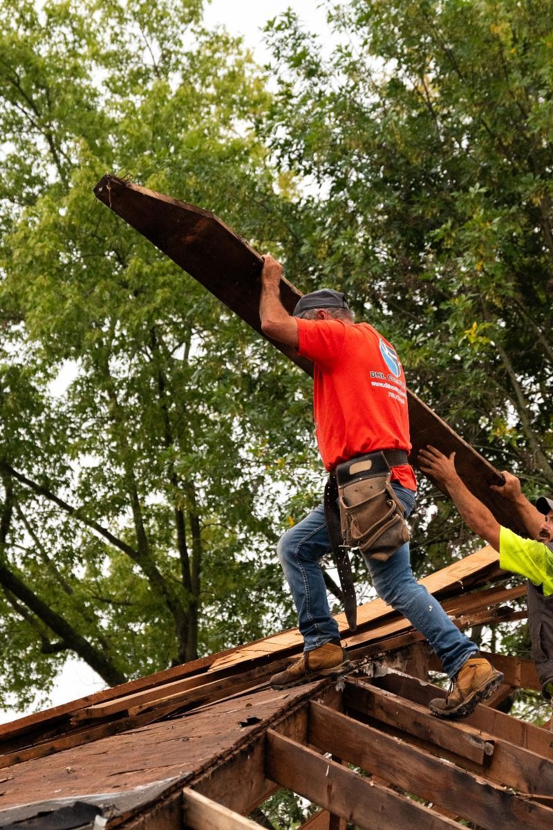 Two construction workers carrying a long, wooden plank on a roof, surrounded by trees.