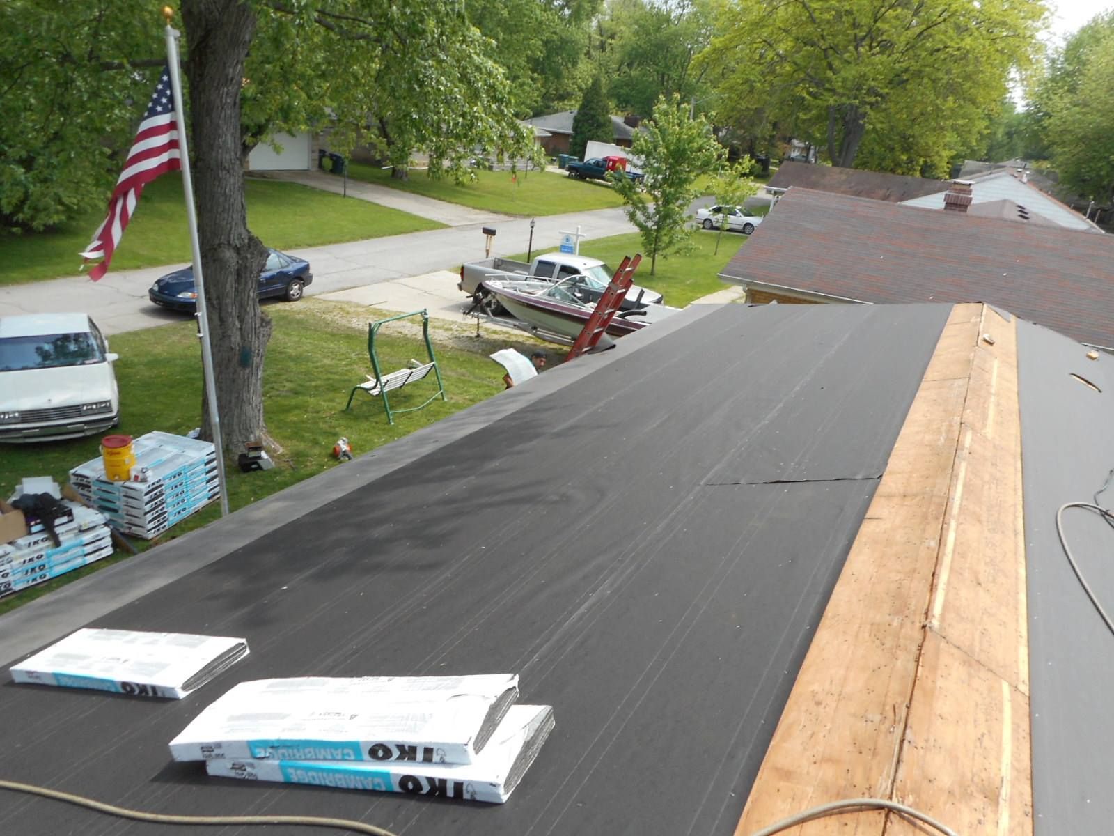 Flat roof with black asphalt, lumber, and stacks of roofing material, with a residential neighborhood in the background.