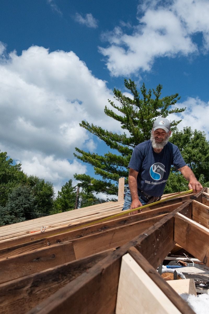 Man on rooftop measuring with a tape measure, blue sky and trees in the background.