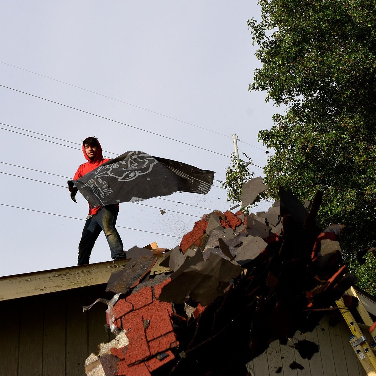 Man on damaged roof holding roofing material. Debris visible; green trees and power lines in the background.