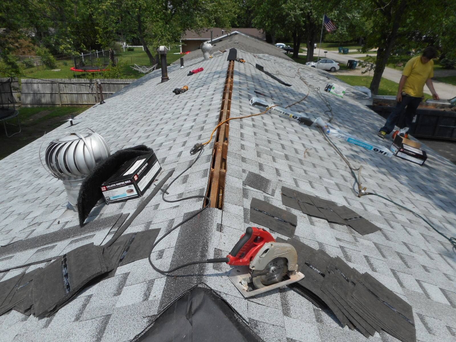 Roofer on a gray asphalt shingle roof, using a circular saw near a vent and other tools. Sunny day.