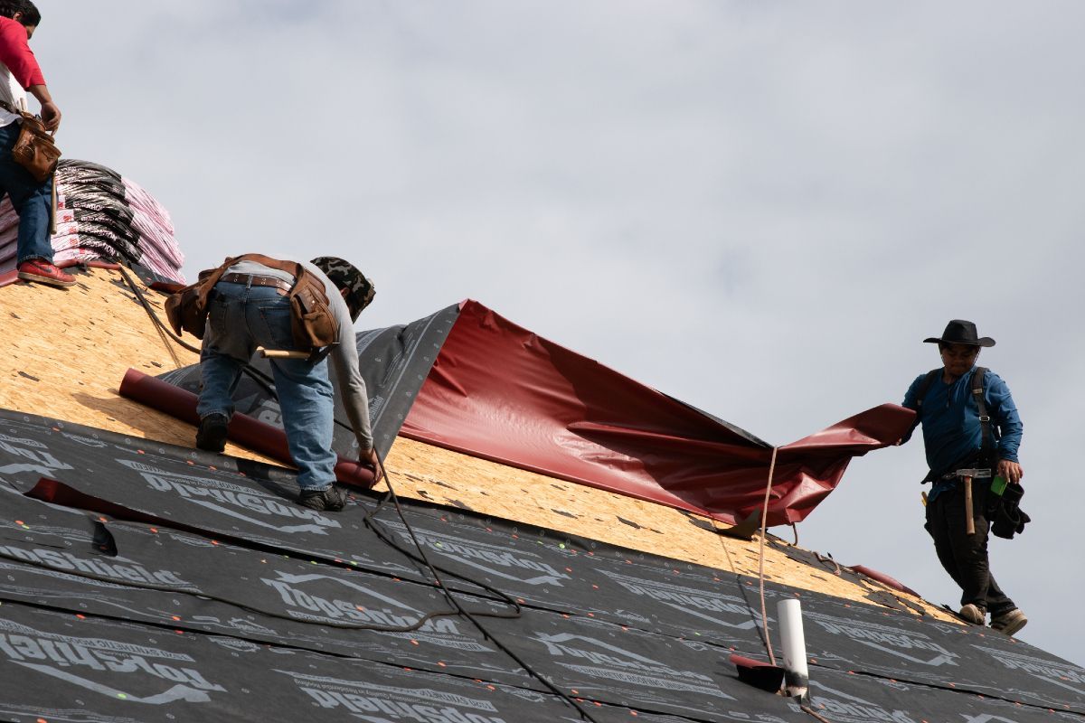 Roofers installing a red underlayment layer on a roof, sunny day.