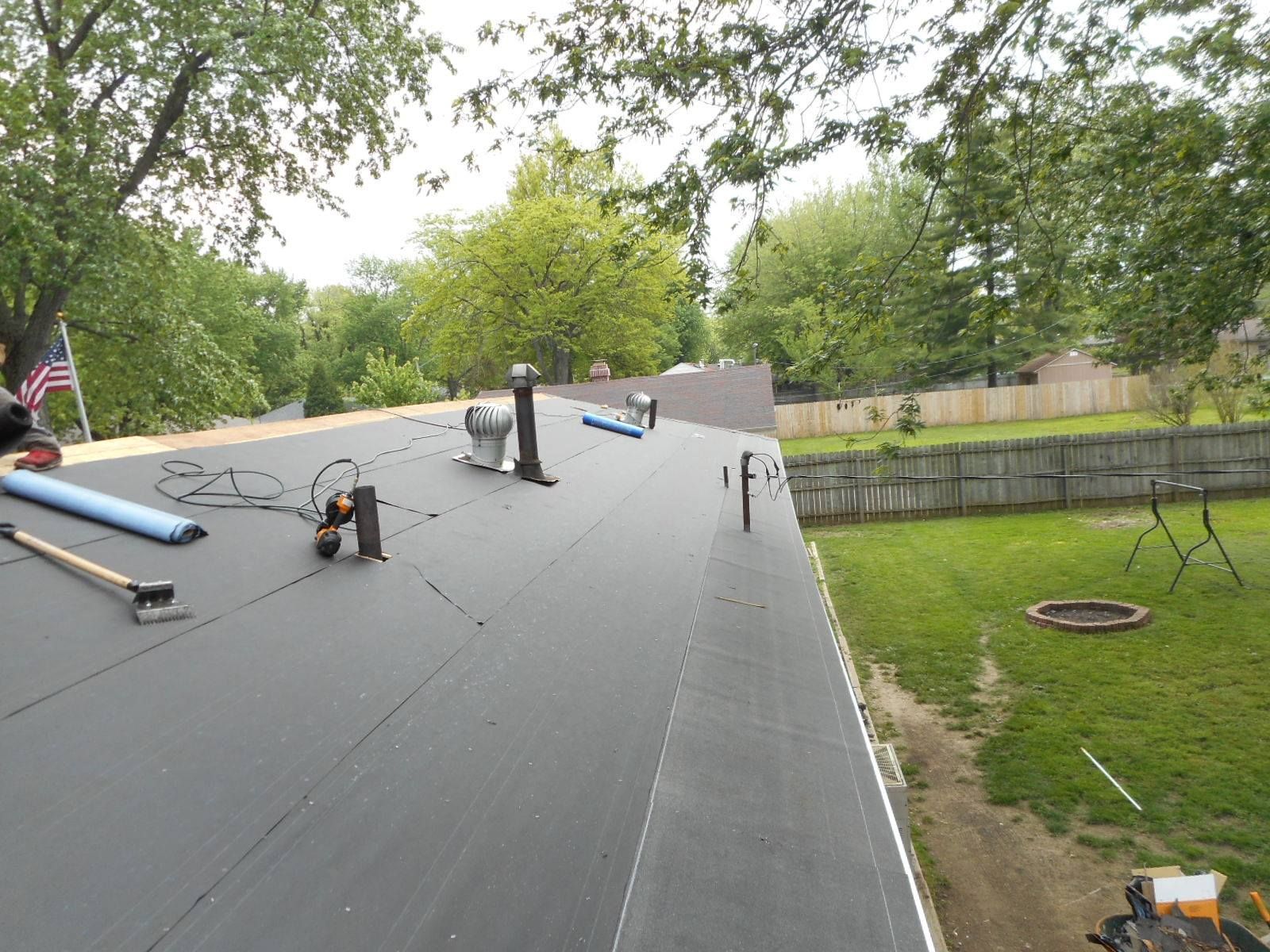 A black roof with tools and vents, overlooking a green backyard with a fence.
