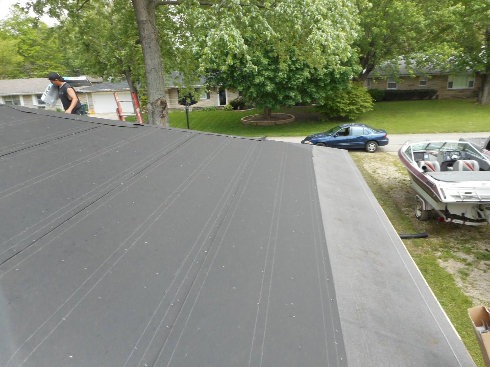 Person on a dark roof, near a tree, working on roofing project in a suburban setting. Car and boat visible.
