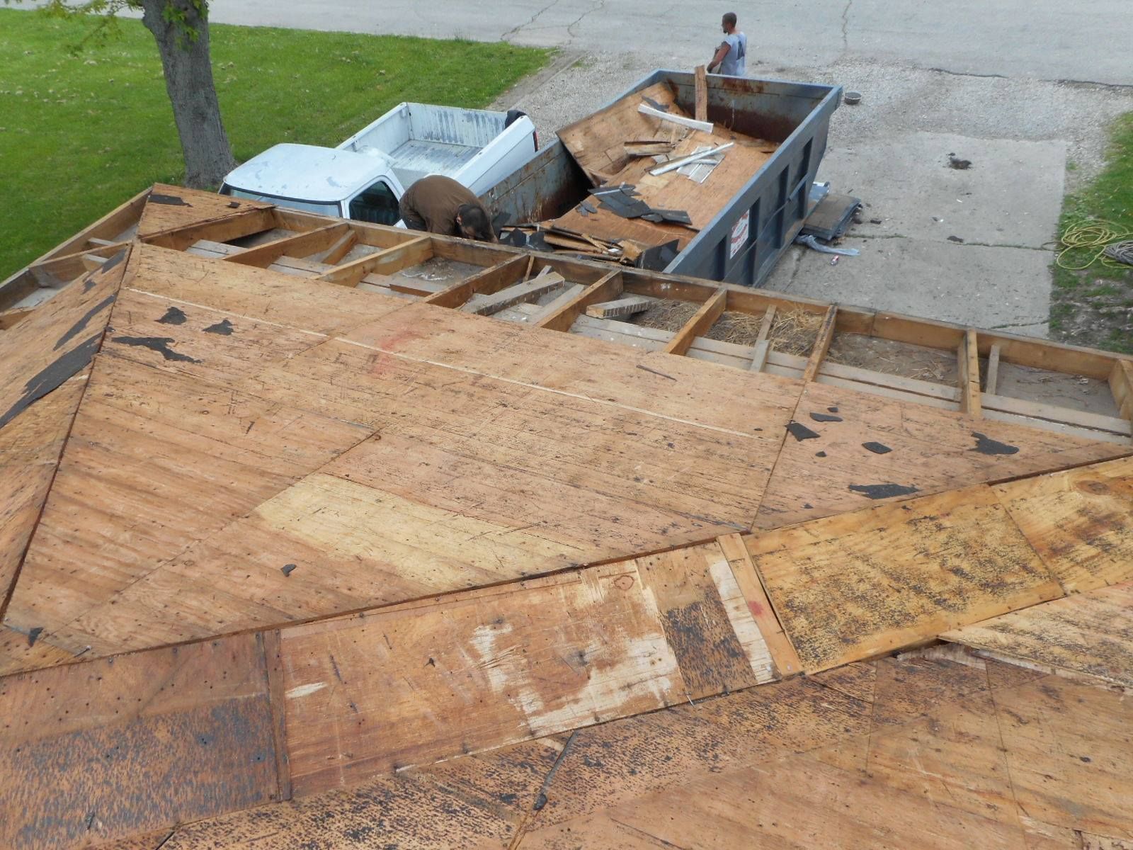 Roof partially stripped; debris in a dumpster next to a truck. Person standing by the dumpster.