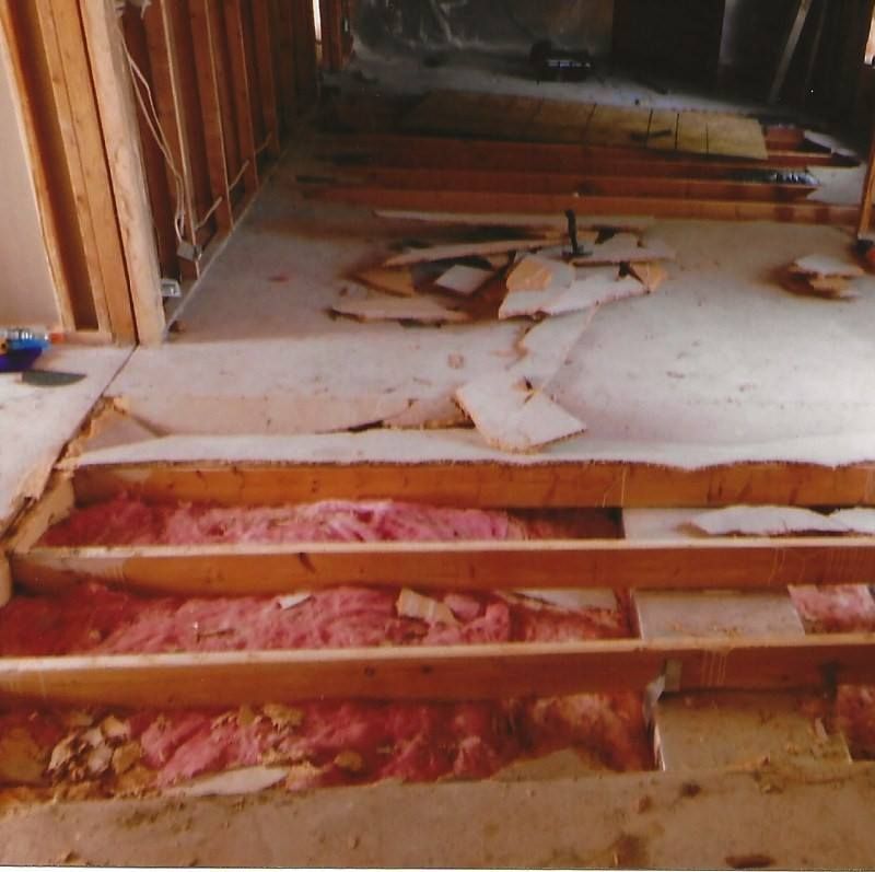 Construction site: exposed floor joists with pink insulation. Rough-framed walls in a room. Debris on the floor.