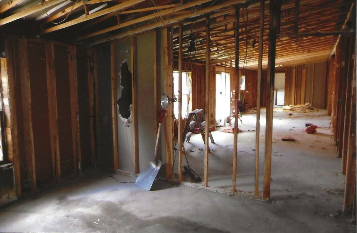 Interior of a house under renovation. Bare studs, exposed beams, and debris scattered on the concrete floor.