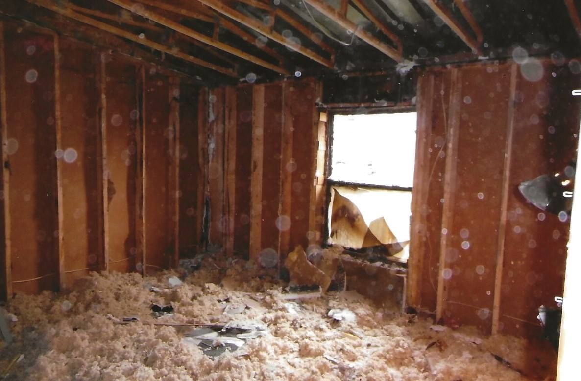 Interior of a damaged room, walls and ceiling charred, window broken, insulation scattered on the floor.