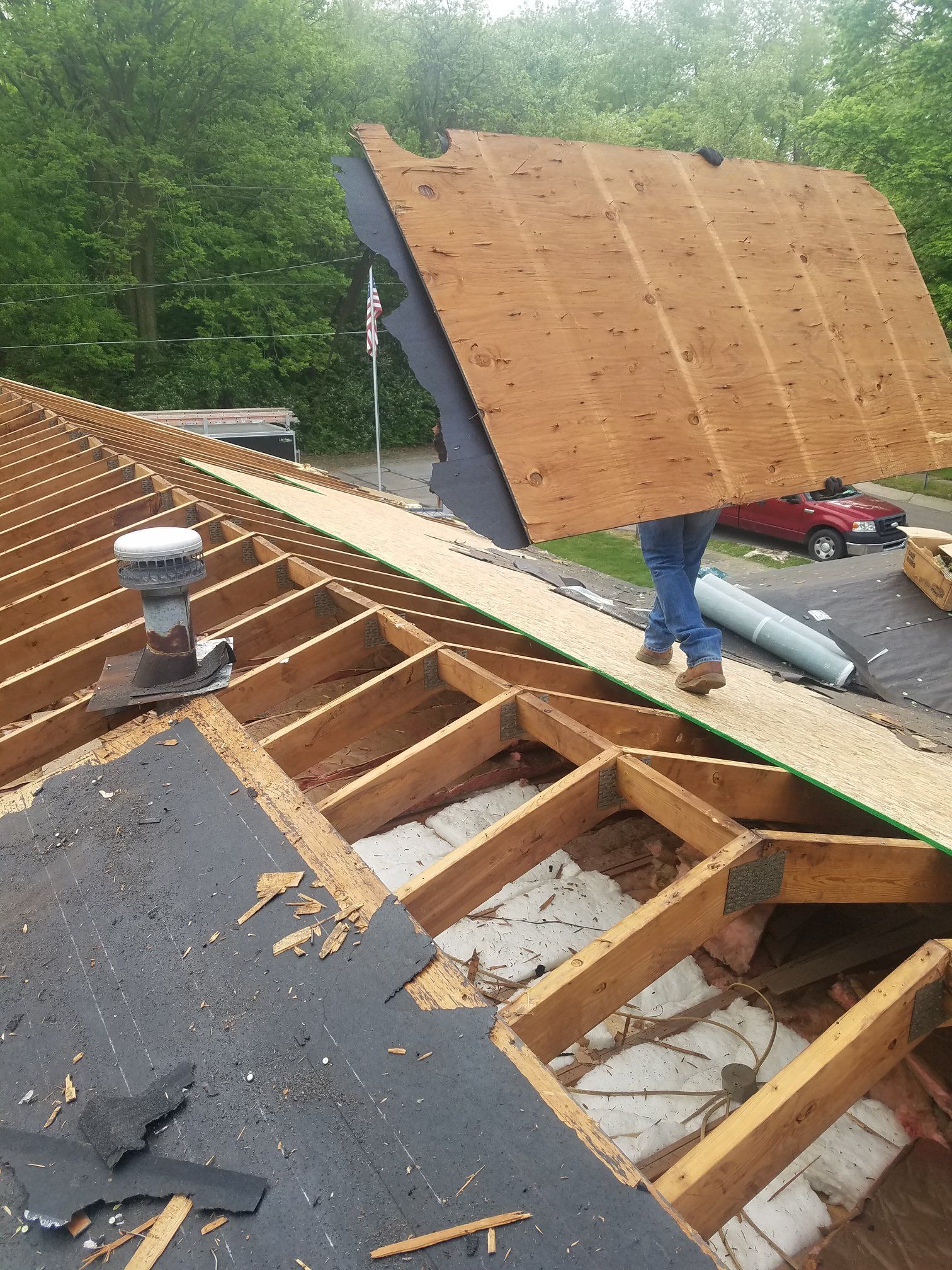 A person carrying a large piece of plywood on a roof during construction. The roof is partially torn off.