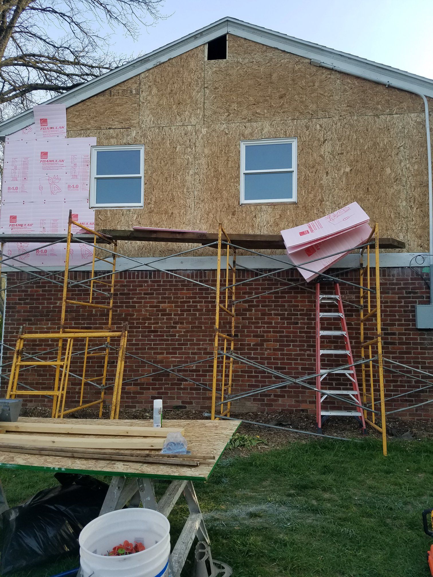 House under construction, pink insulation being installed over brick and wood siding. Scaffolding and ladder present.