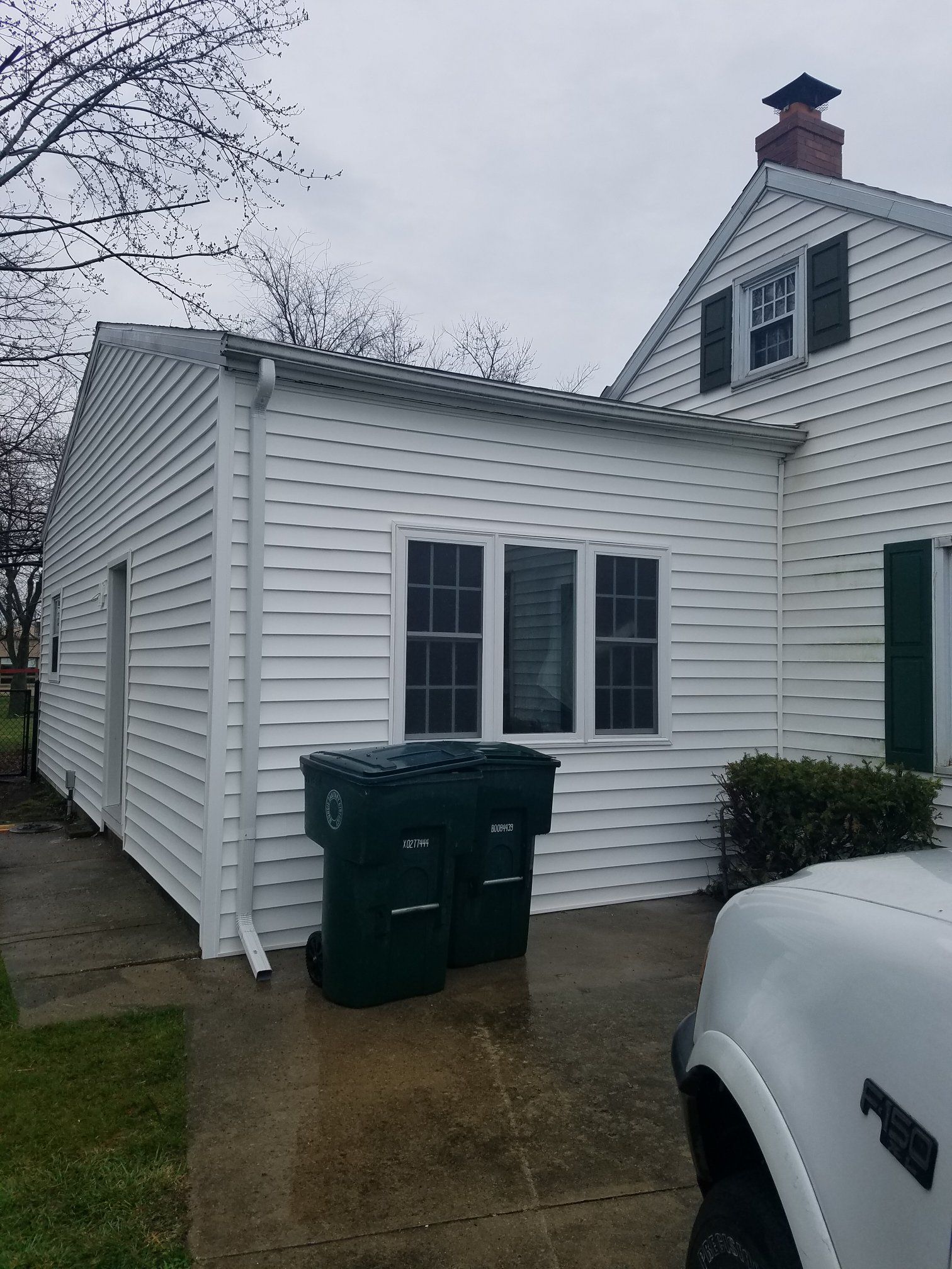White house with a side addition, two windows, two green trash cans. Overcast day.