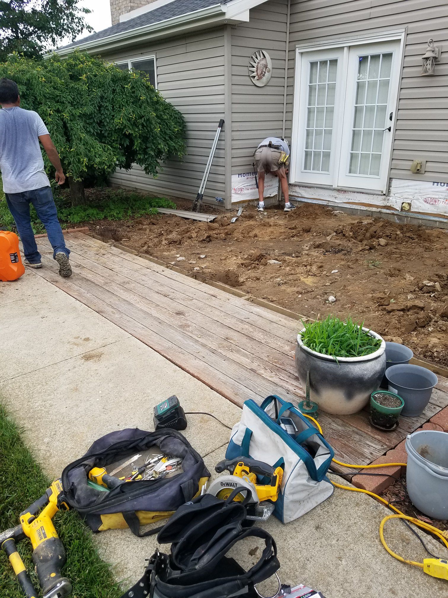 Workers near a house removing ground material. Tools and a potted plant are visible nearby.