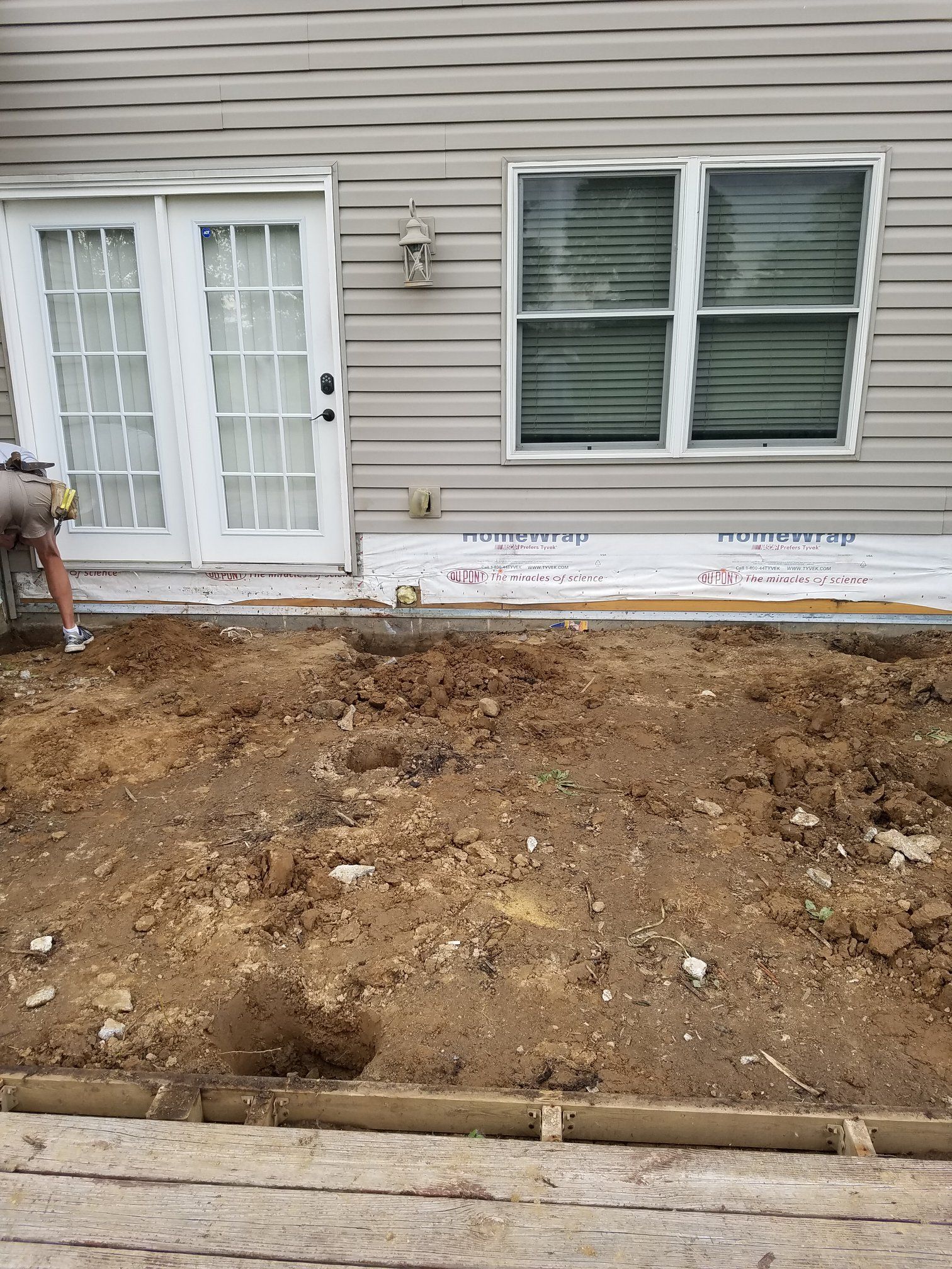 Exterior view of a house with French doors and a window. Excavated dirt area near the foundation. Boards laid out.