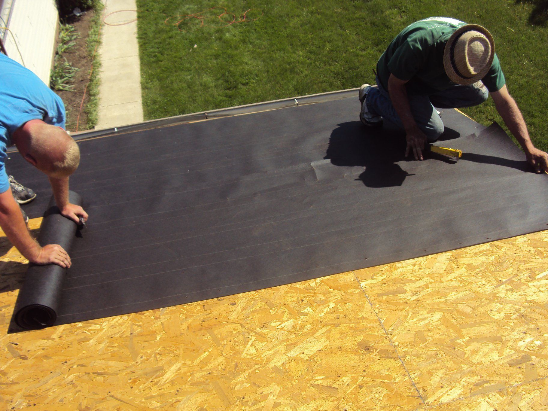 Two people installing black roofing underlayment on an OSB roof deck.