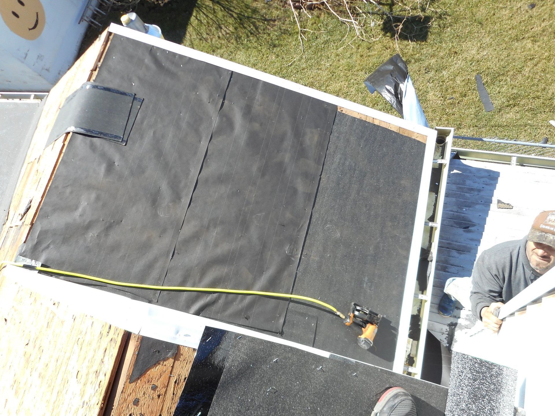 Roofer installing black roofing material on a small roof section, using a nail gun.