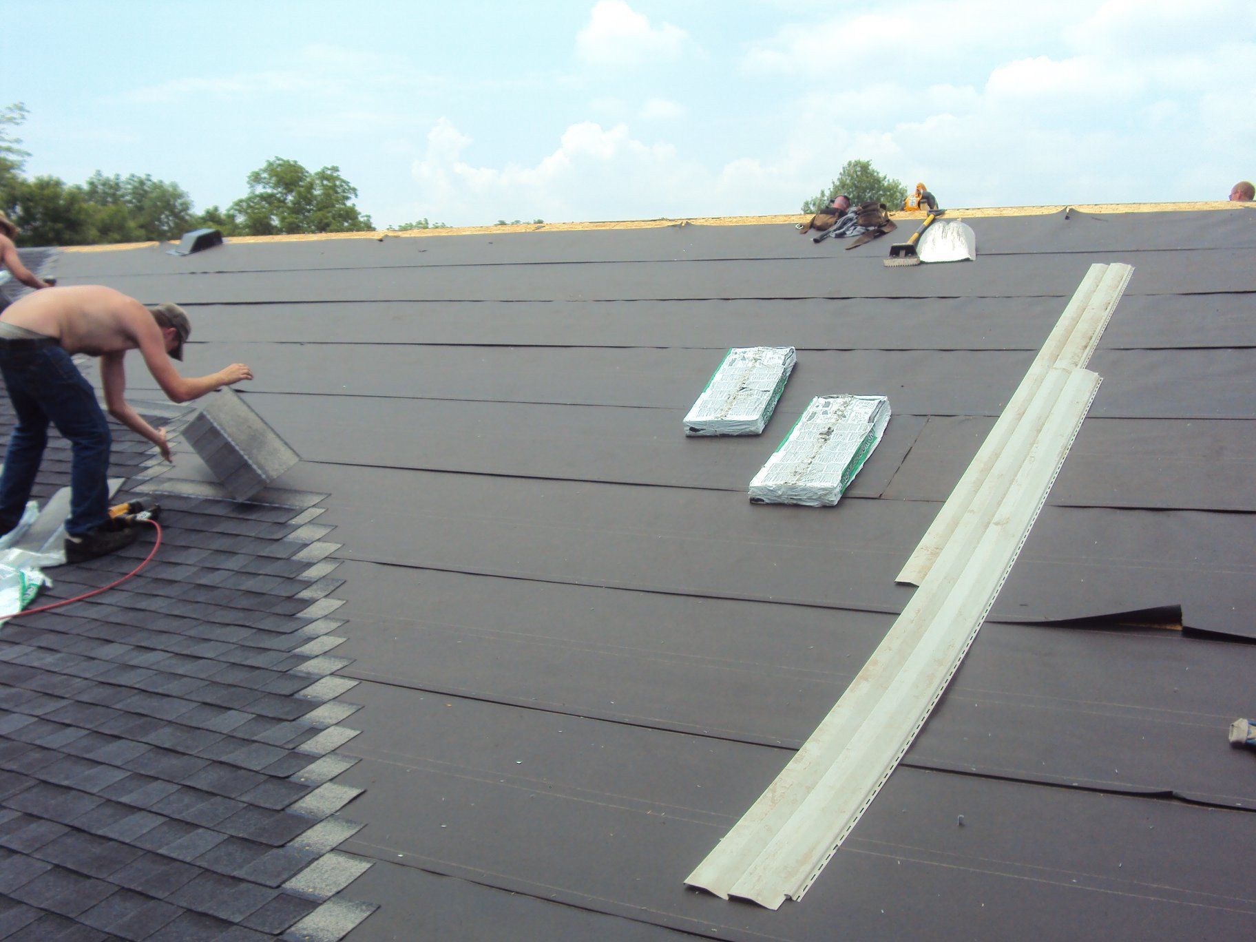 Workers on a roof installing shingles. One shirtless man working with metal flashing. New black underlayment is visible.