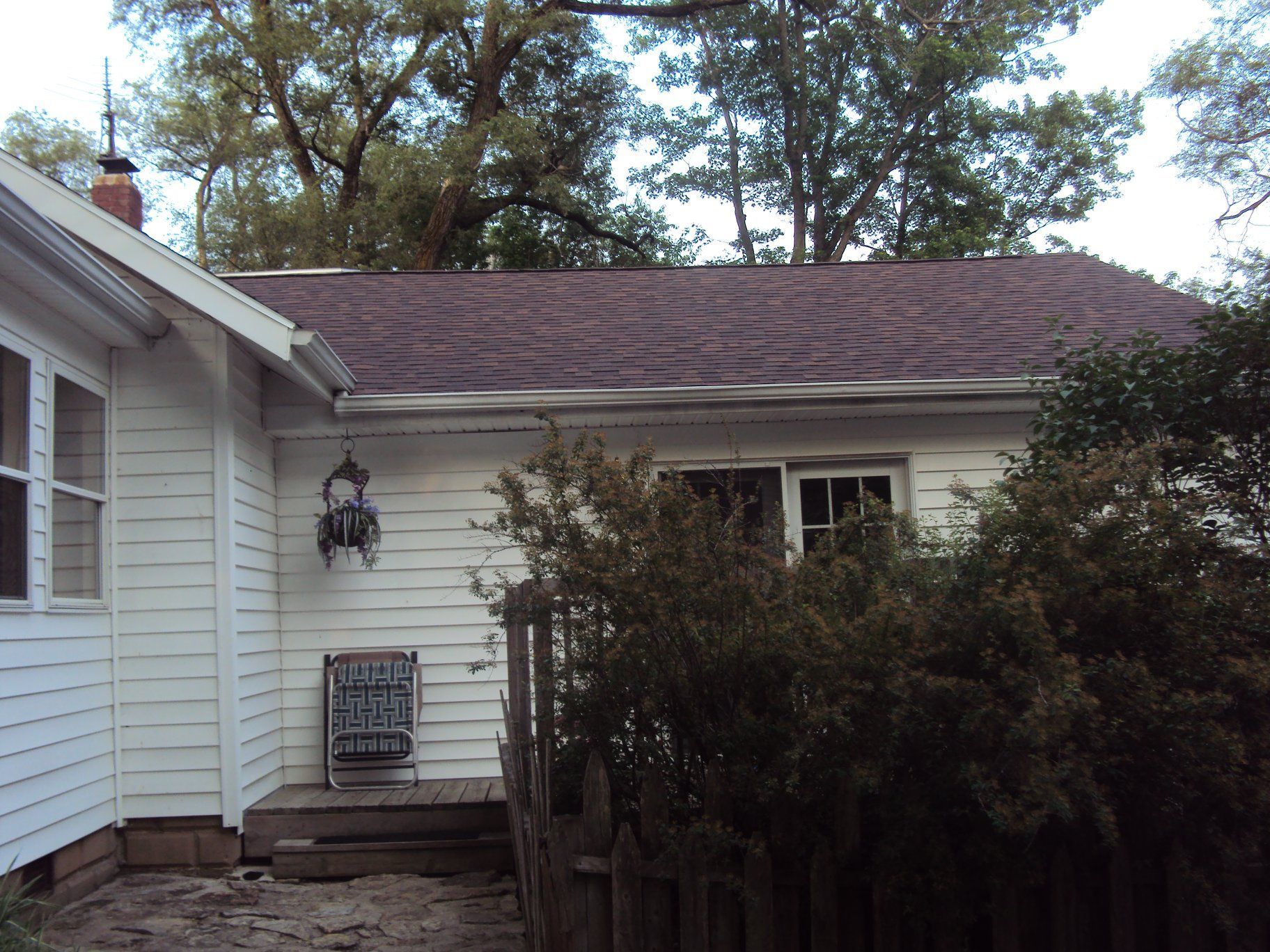 White house exterior with dark brown roof, trees in the background, a small porch, and overgrown bushes.