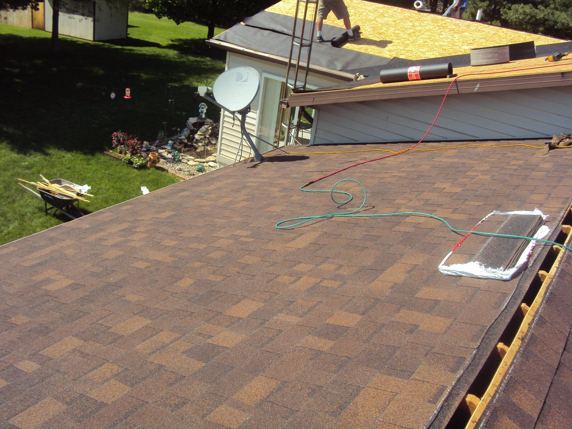 Roofer working on a brown shingle roof; satellite dish and ladder present. Green yard in the background.