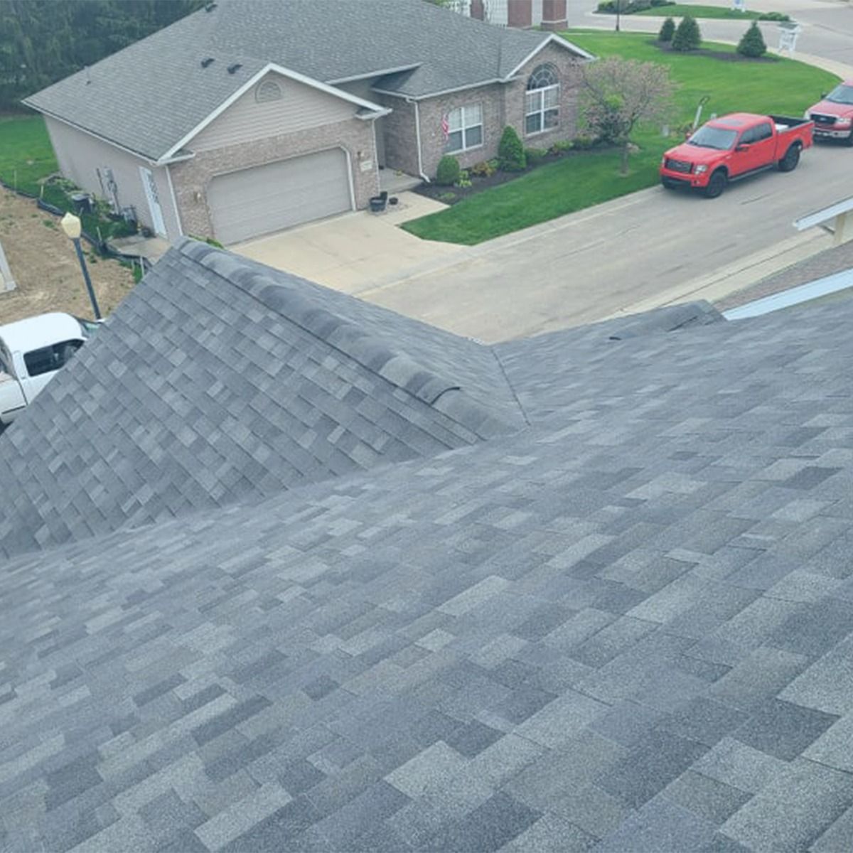 Gray asphalt shingle roof, angled view; houses, red trucks, and green lawn in background.