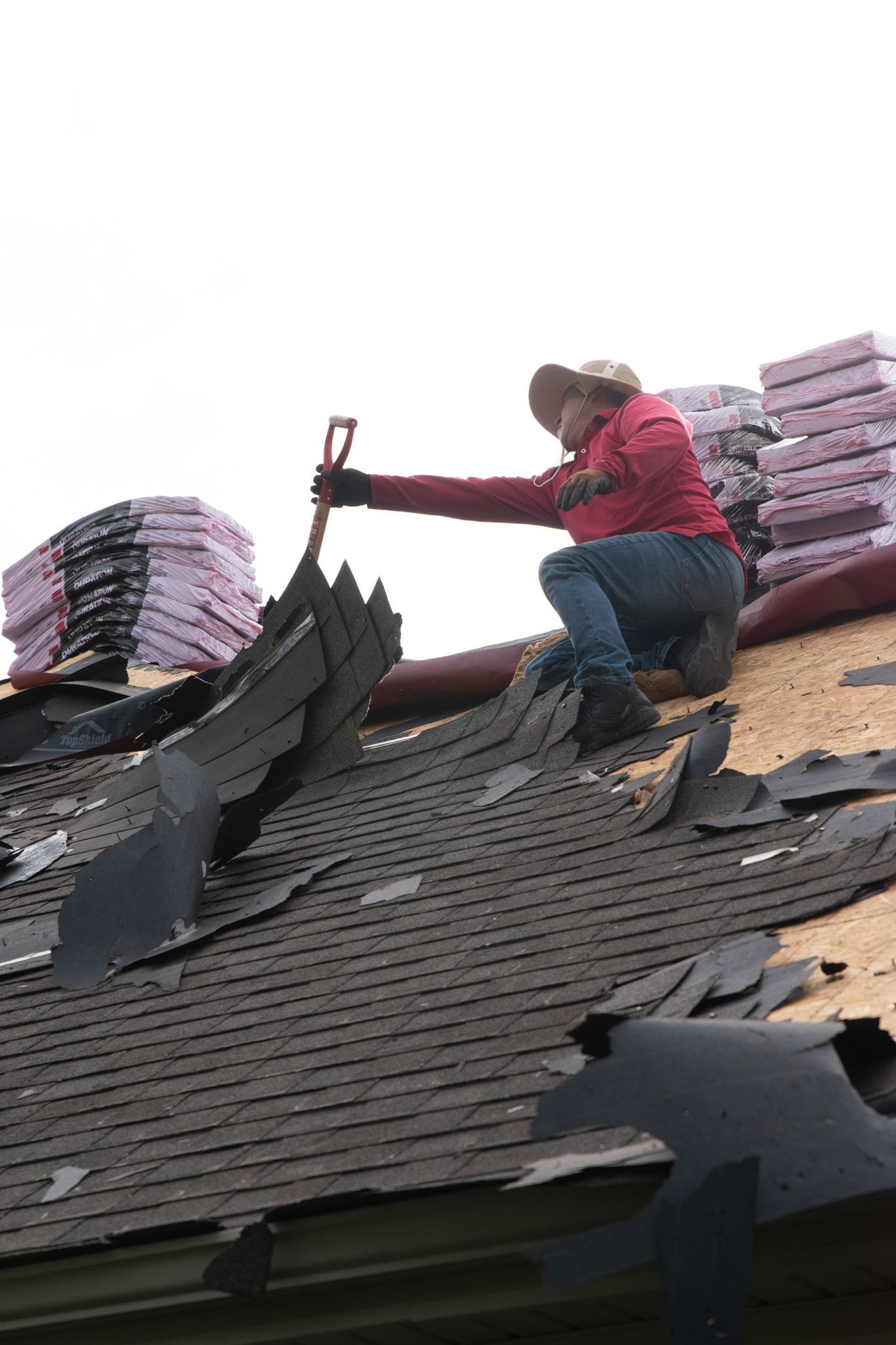 Roofer removes old shingles with a shovel on a sloped roof.  Pink insulation and new shingles stacked in background.