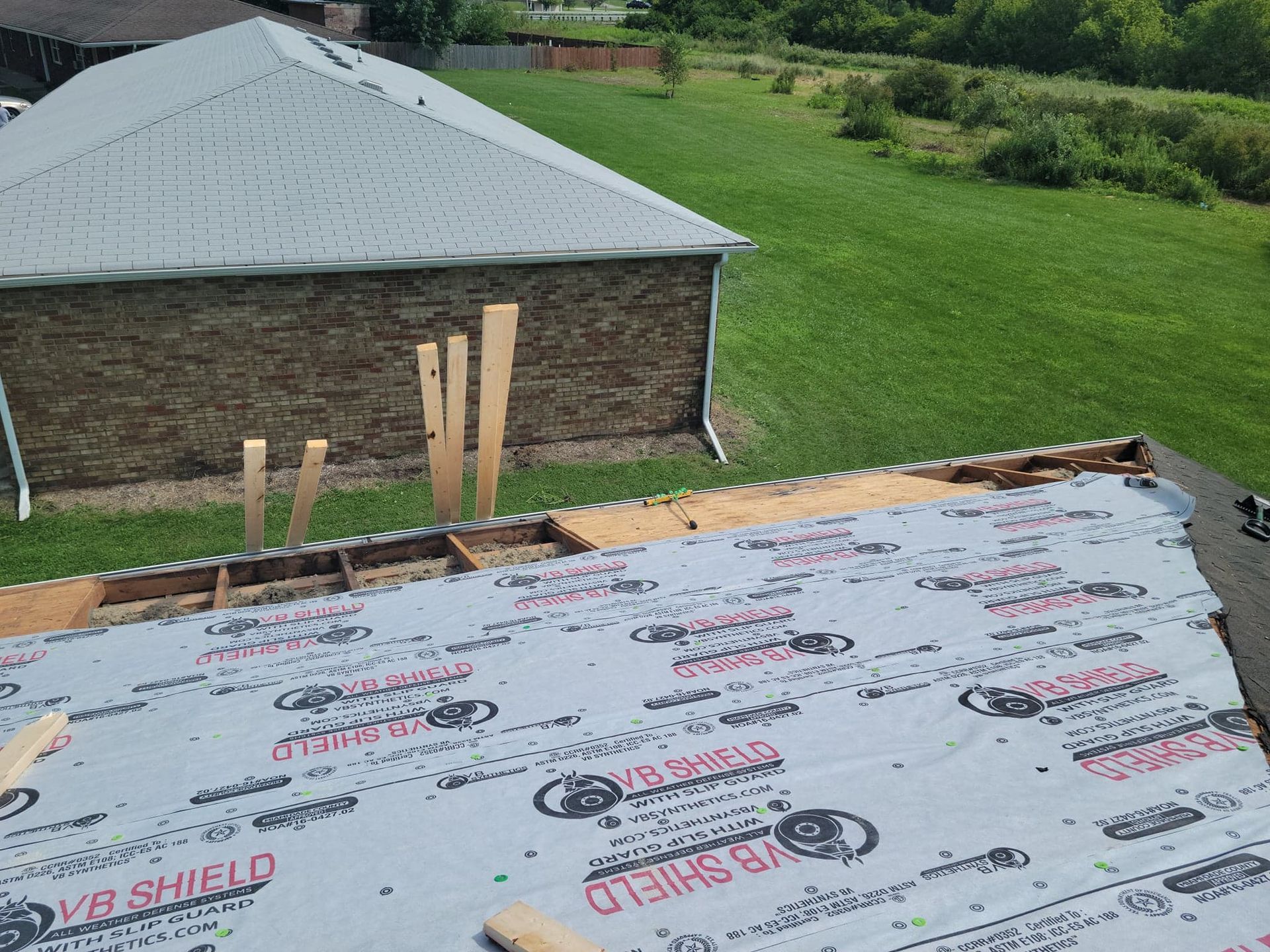 Roof being worked on, plywood and protective underlayment visible, with building and green yard in background.