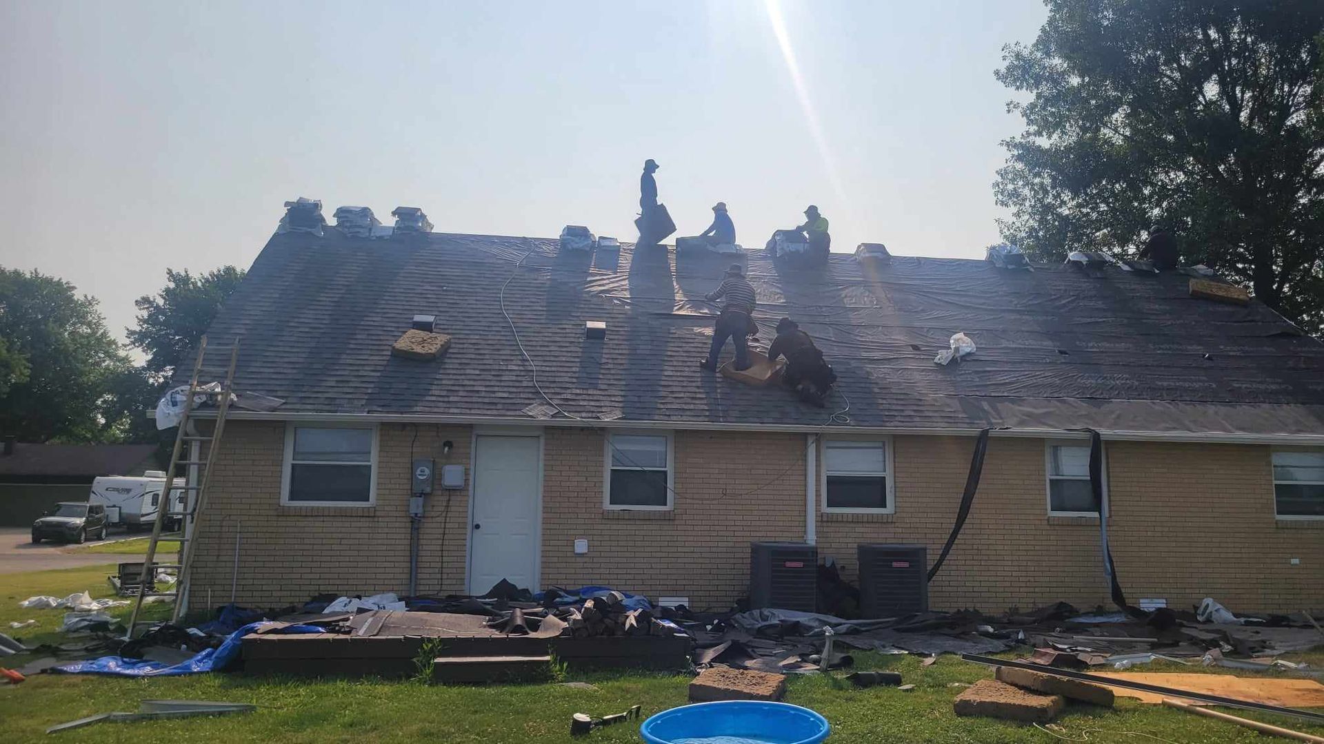 Workers on a house roof, removing shingles. Blue tarp and debris on the ground. Sunny day.
