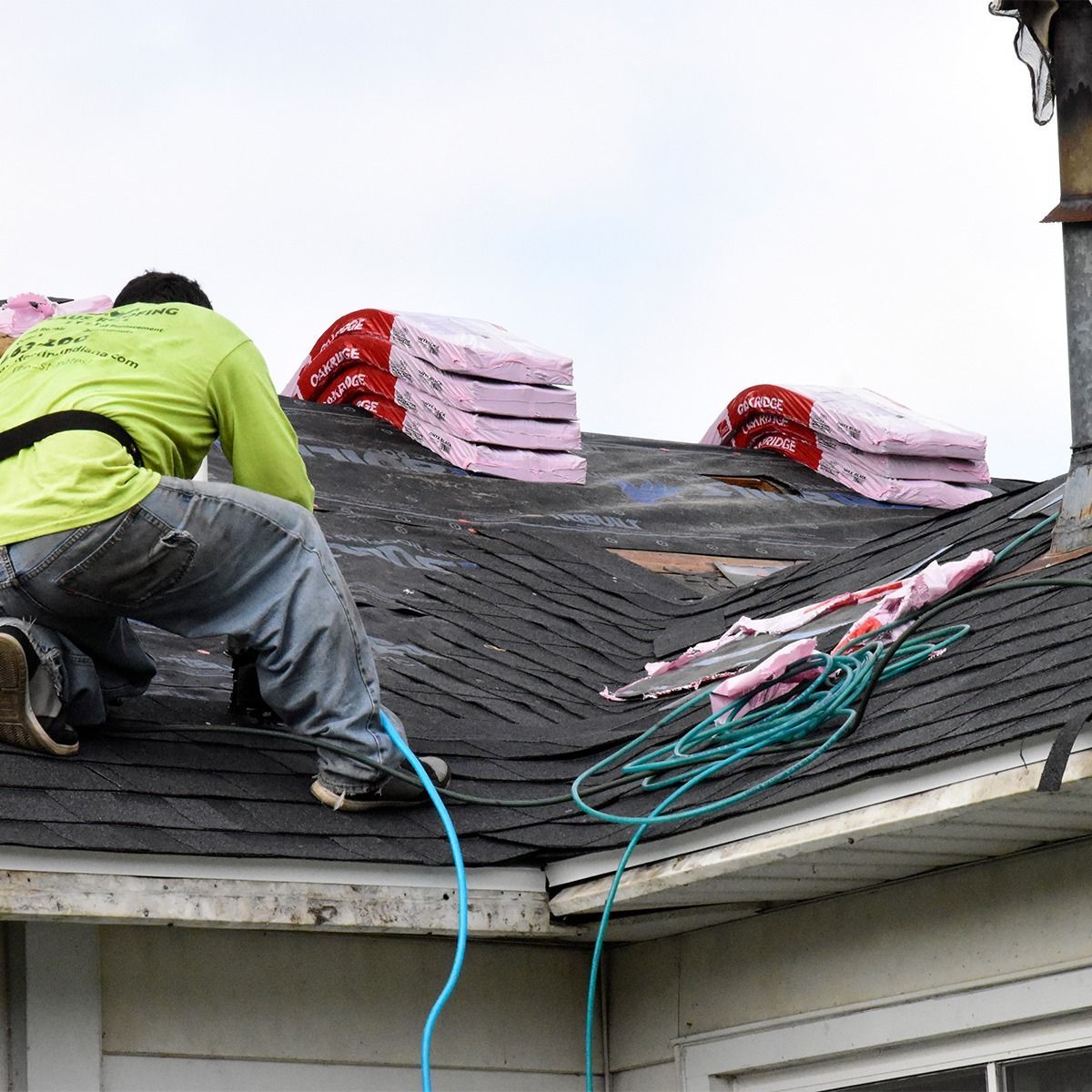 Roofer in green shirt works on a roof, surrounded by bundles of pink insulation and exposed underlayment.
